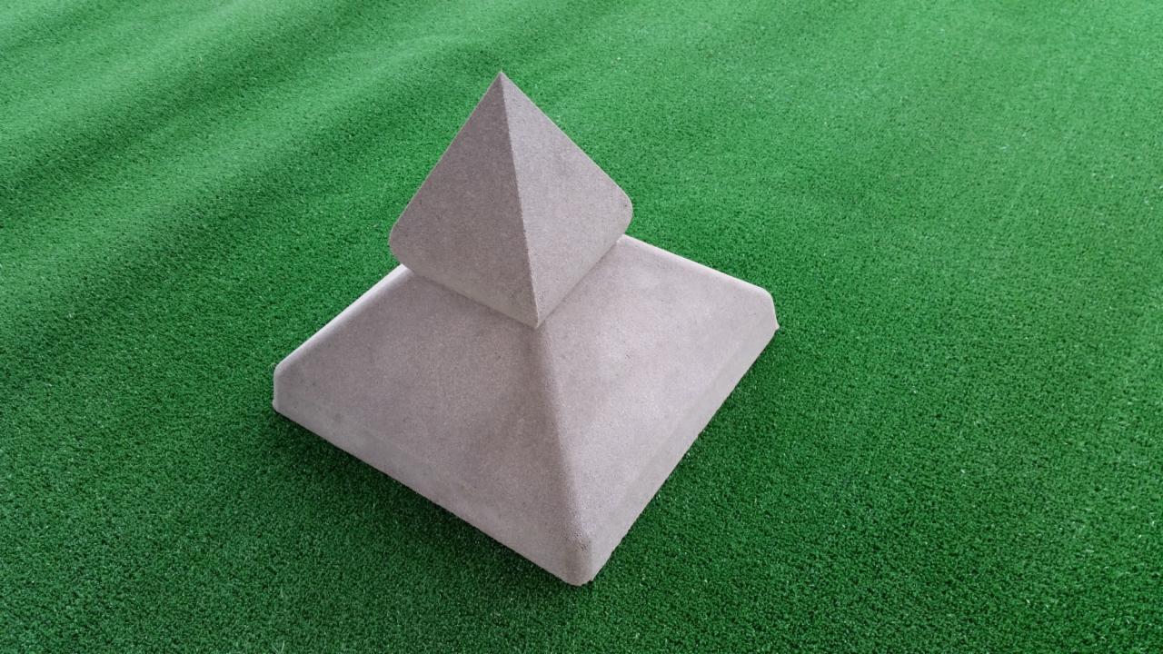 Szary, betonowy słupek ogrodzeniowy z piramidalnym zwieńczeniem na tle sztucznej, zielonej trawy.