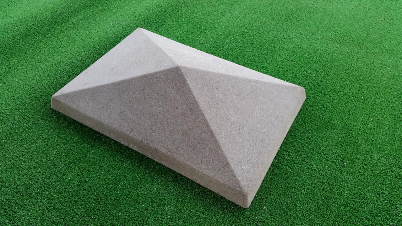 Betonowy słupek ogrodzeniowy z piramidalnym zwieńczeniem, leżący na sztucznej trawie.