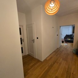 Formela Maciej Abcwnetrza - Jasne wnętrze po odświeżeniu: białe ściany, drewniana podłoga, nowoczesna lampa wisząca. Widoczne drzwi i fragment pokoju w głębi. Minimalistyczny design.