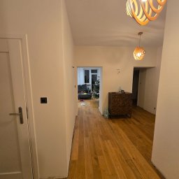 Formela Maciej Abcwnetrza - Jasne wnętrze po malowaniu: parkiet, białe ściany, drzwi, designerskie lampy. W tle widoczny pokój z choinką. Minimalistyczny styl.