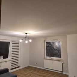 Formela Maciej Abcwnetrza - Jasne wnętrze pokoju po remoncie: świeżo pomalowane ściany w odcieniach bieli, drewniana podłoga, okna zabezpieczone folią malarską, nowoczesna lampa sufitowa.