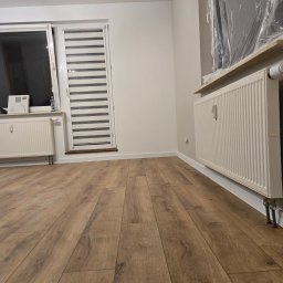 Formela Maciej Abcwnetrza - Jasne panele podłogowe w nowym pokoju z grzejnikami i oknem z żaluzjami. Ściany pomalowane na biało, jeden grzejnik zabezpieczony folią malarską.