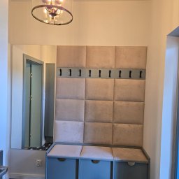 Formela Maciej Abcwnetrza - Stylowy przedpokój z tapicerowanym panelem ściennym, wieszakami i szafką z siedziskiem w odcieniach beżu i szarości. Dekoracyjna lampa i wzorzysta podłoga.