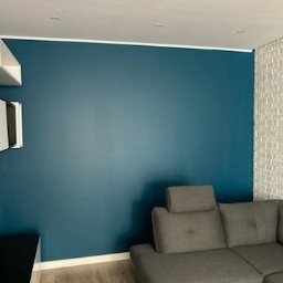 Formela Maciej Abcwnetrza - Ściana pomalowana na ciemny turkus w salonie z szarą sofą i fragmentem ściany z białej cegły. Widoczne oświetlenie sufitowe i drewniana podłoga.