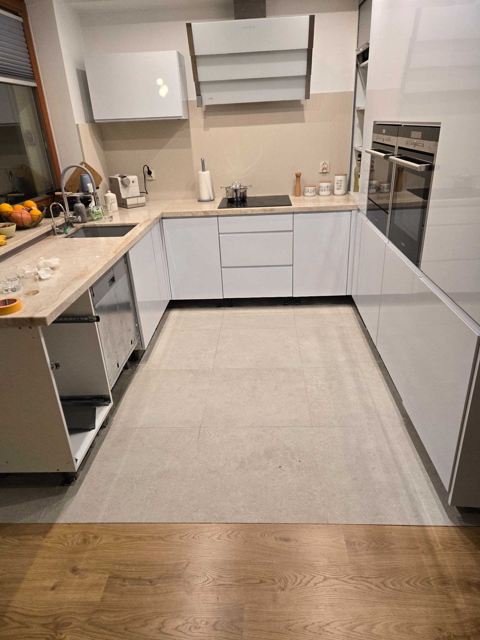 Nowoczesna kuchnia w trakcie montażu: białe szafki, marmurowy blat, widoczne niedokończone elementy, jasna podłoga. Ujęcie z perspektywy podłogi.