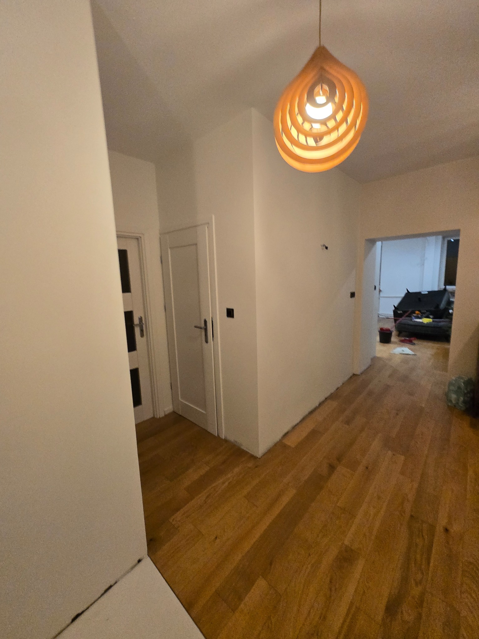 Jasne wnętrze po odświeżeniu: białe ściany, drewniana podłoga, nowoczesna lampa wisząca. Widoczne drzwi i fragment pokoju w głębi. Minimalistyczny design.