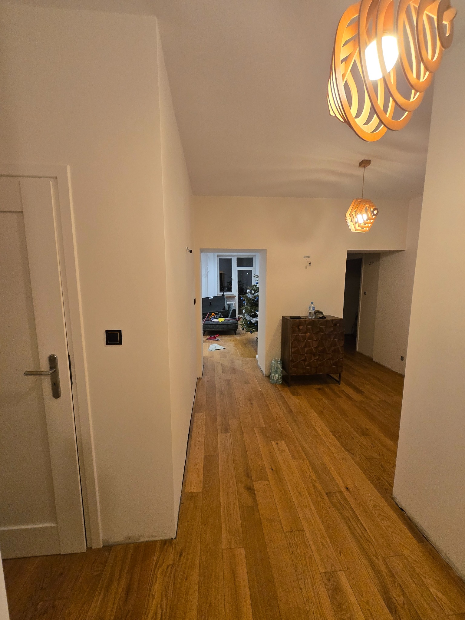 Jasne wnętrze po malowaniu: parkiet, białe ściany, drzwi, designerskie lampy. W tle widoczny pokój z choinką. Minimalistyczny styl.