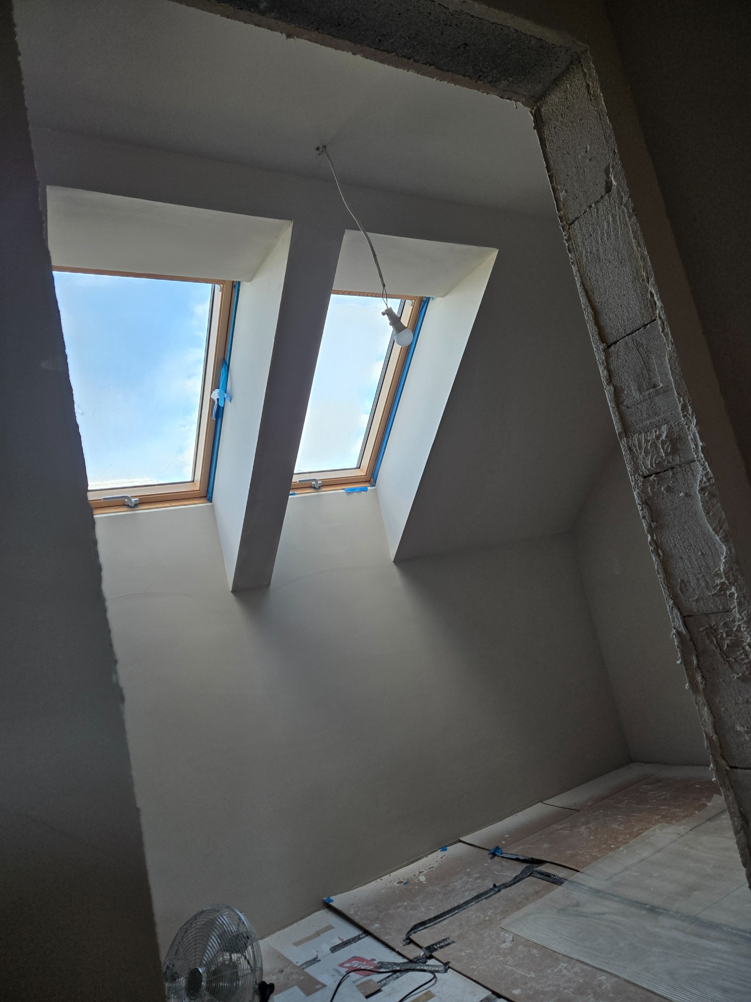 Poddasze po gładziowaniu ścian i sufitu, widoczne okna dachowe zabezpieczone taśmą malarską, wentylator podłogowy i surowa ościeżnica. Prace wykończeniowe w toku.