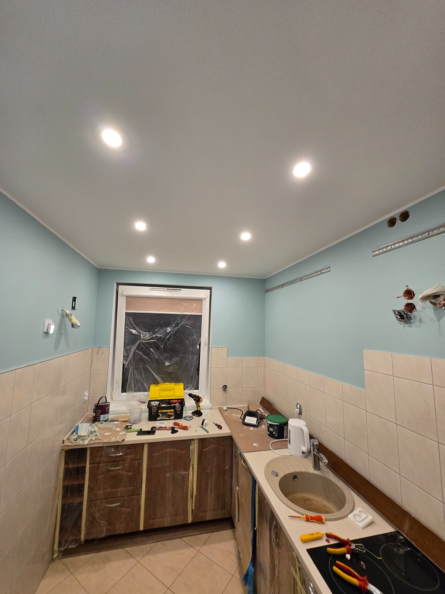 Kuchnia w trakcie remontu: błękitne ściany, częściowo wyłożone kafelkami, okno zabezpieczone folią, widoczne narzędzia i akcesoria, przygotowane do dalszych prac wykończeniowych.