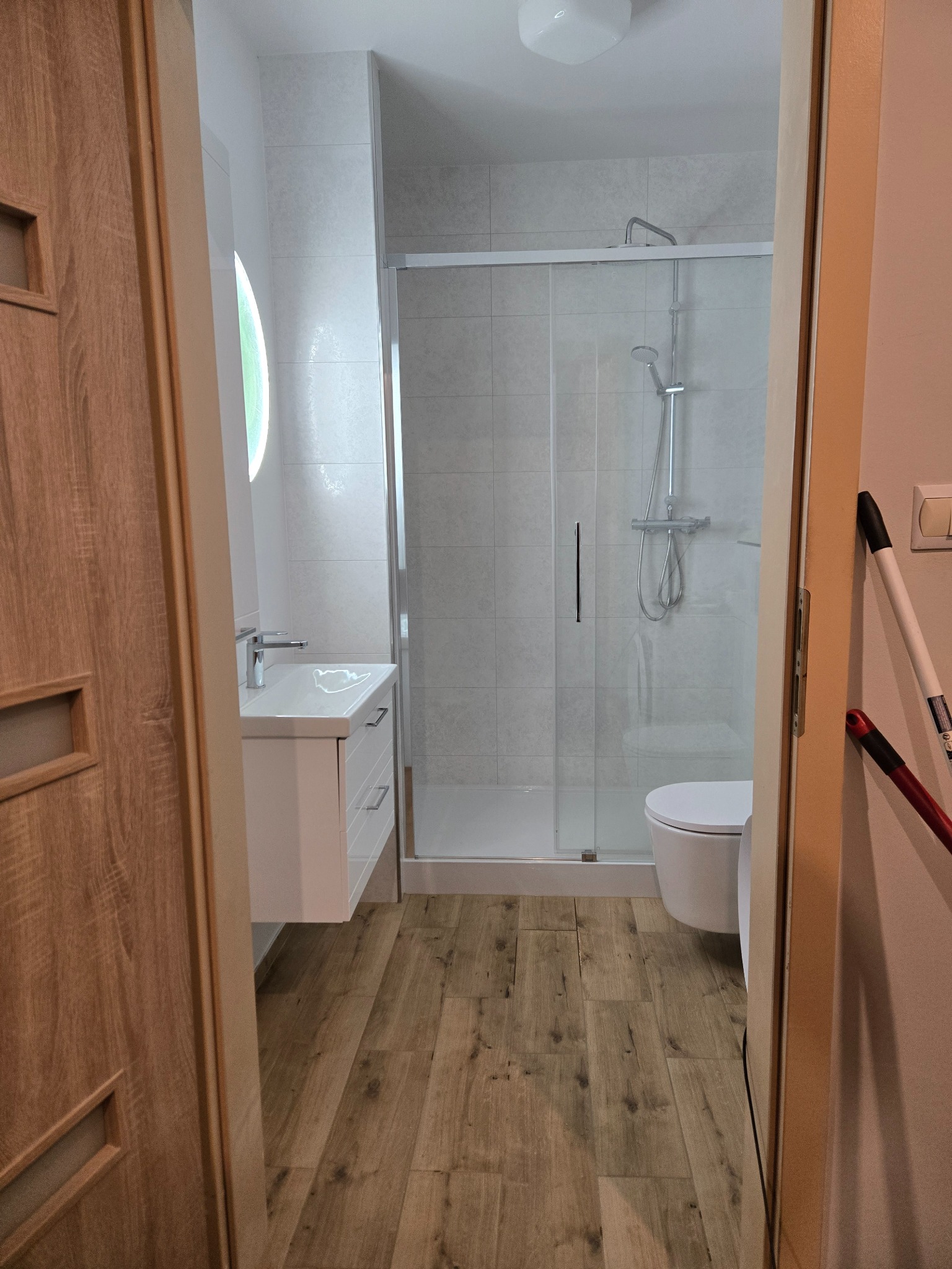 Nowoczesna łazienka z kabiną prysznicową, umywalką z szafką i toaletą. Jasne płytki i drewnopodobna podłoga tworzą spójną całość. Widok zza uchylonych drzwi.