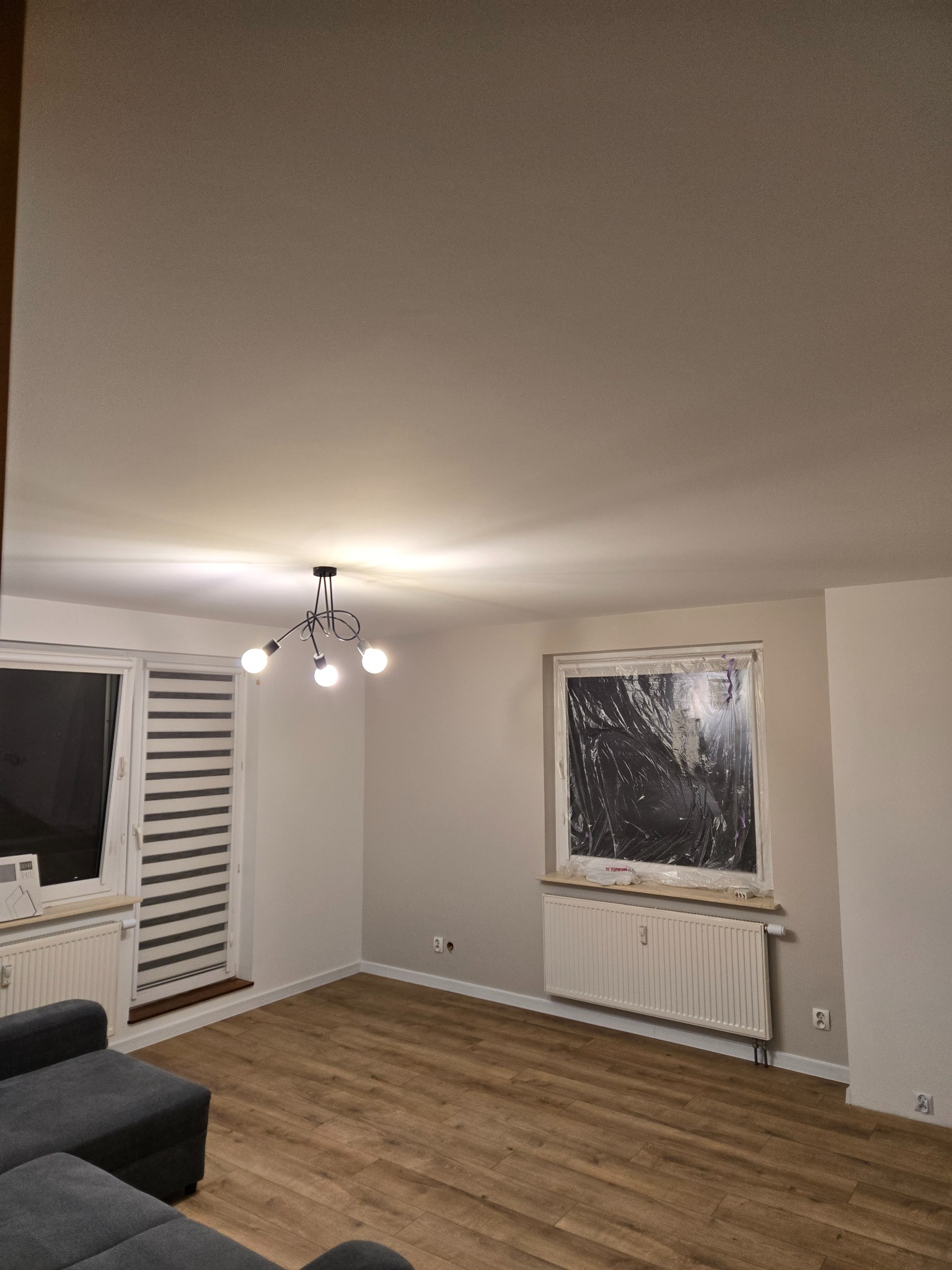 Jasne wnętrze pokoju po remoncie: świeżo pomalowane ściany w odcieniach bieli, drewniana podłoga, okna zabezpieczone folią malarską, nowoczesna lampa sufitowa.