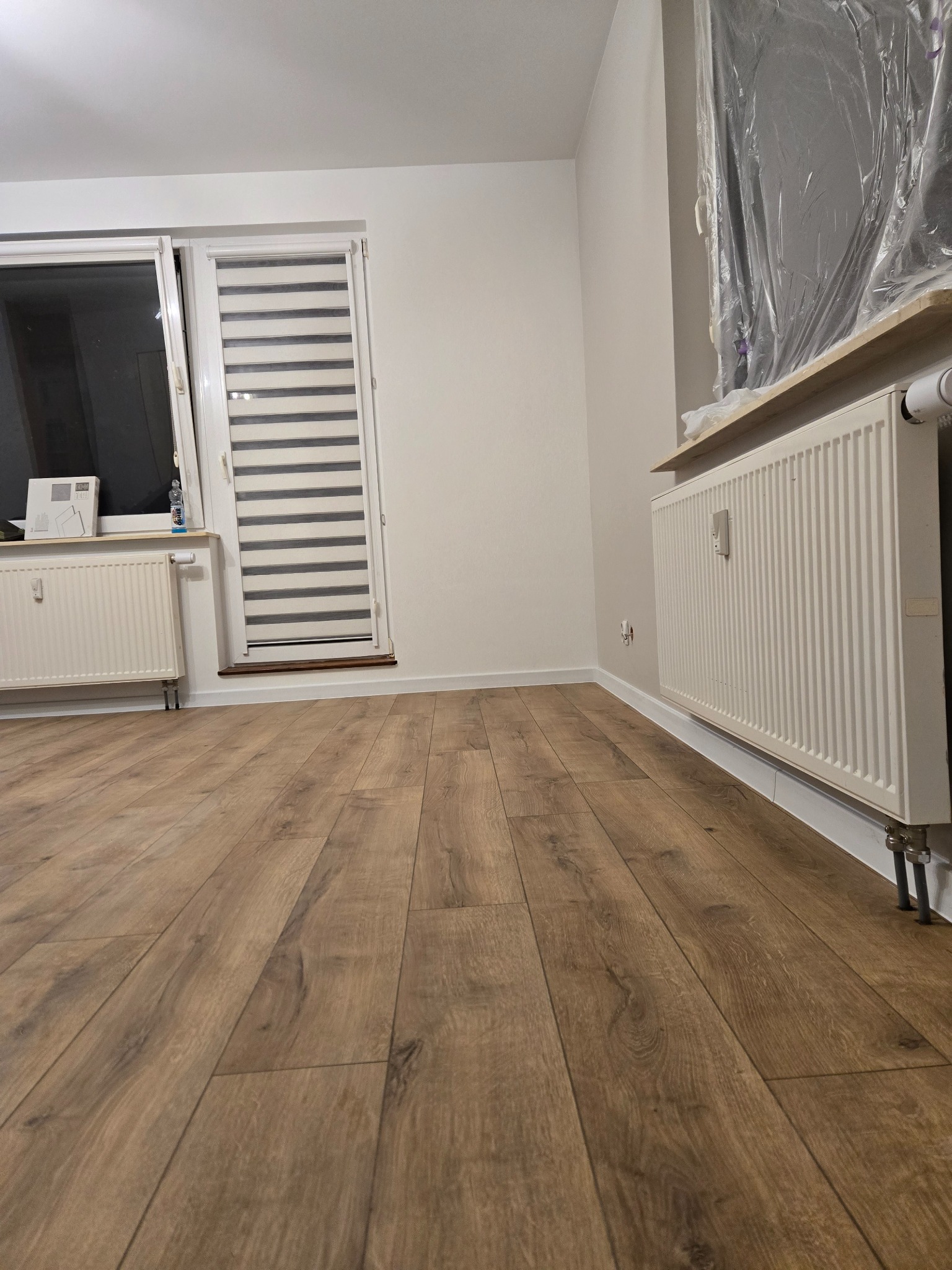 Jasne panele podłogowe w nowym pokoju z grzejnikami i oknem z żaluzjami. Ściany pomalowane na biało, jeden grzejnik zabezpieczony folią malarską.