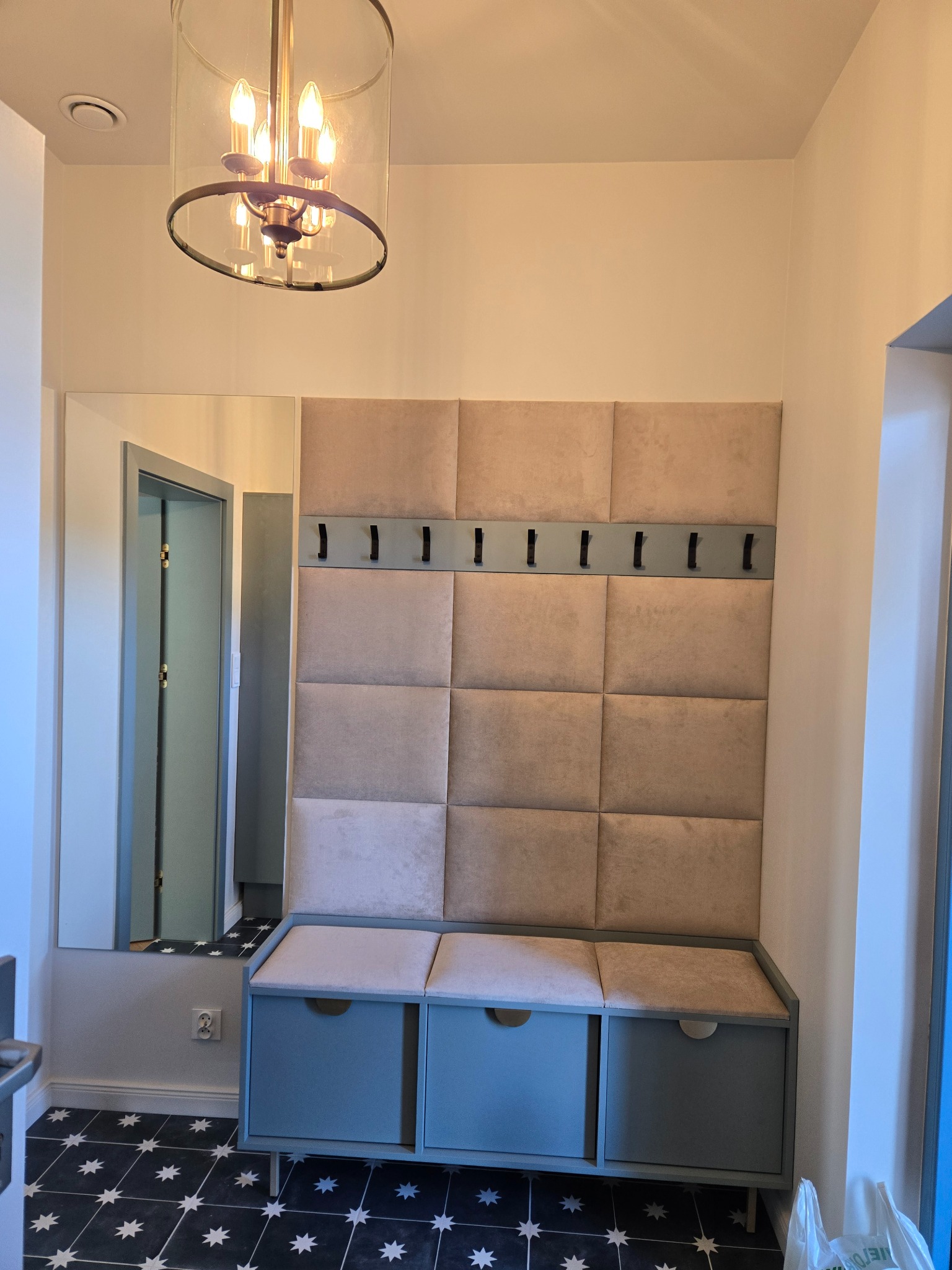 Stylowy przedpokój z tapicerowanym panelem ściennym, wieszakami i szafką z siedziskiem w odcieniach beżu i szarości. Dekoracyjna lampa i wzorzysta podłoga.