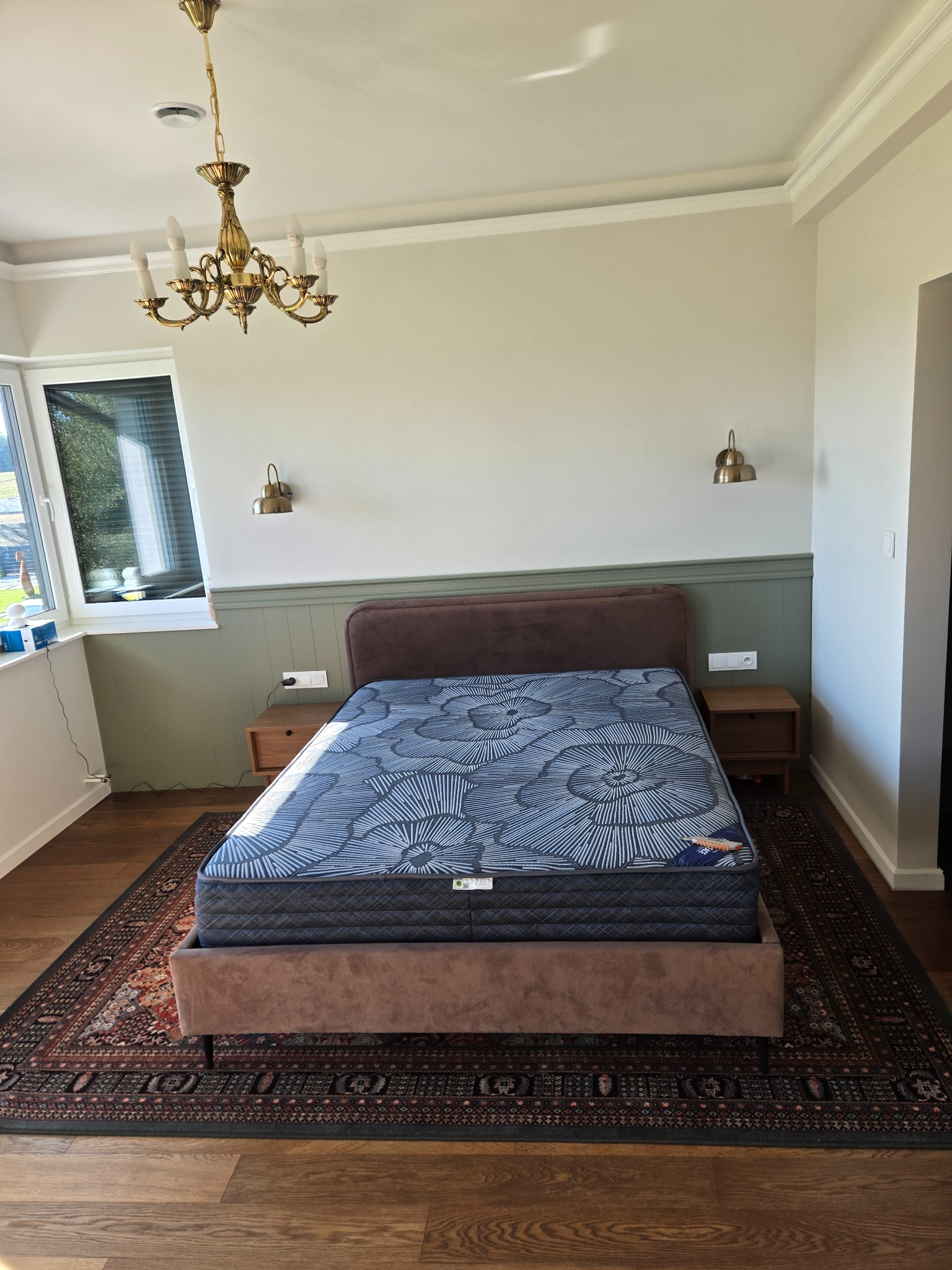 Sypialnia w Gdańsku po wykończeniu: łóżko z brązowym wezgłowiem, wzorzysty materac, drewniana podłoga i dywan w orientalnym stylu. Ściany w odcieniach bieli i zieleni.