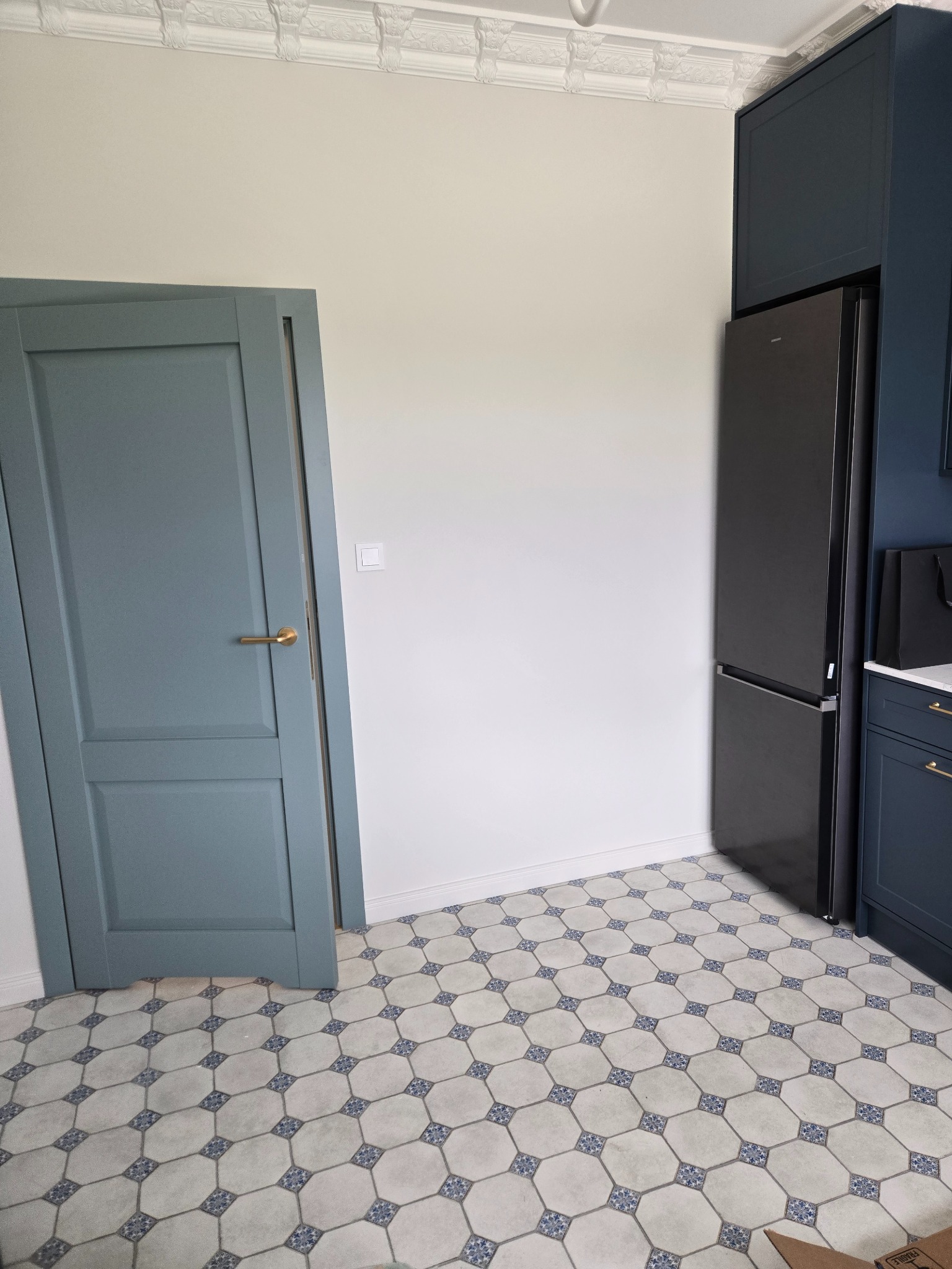 Niebieskie szafki kuchenne i lodówka w nowocześnie wykończonej kuchni z wzorzystą podłogą i ozdobnym gzymsem sufitowym. Widoczny fragment drzwi.