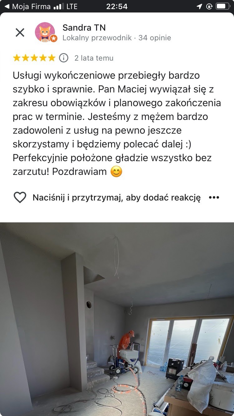 Wnętrze po gładziowaniu ścian, widoczny pracownik z maszyną natryskową. Ściany szare, duże okno z widokiem na zewnątrz. Opinia klienta na górze zdjęcia.