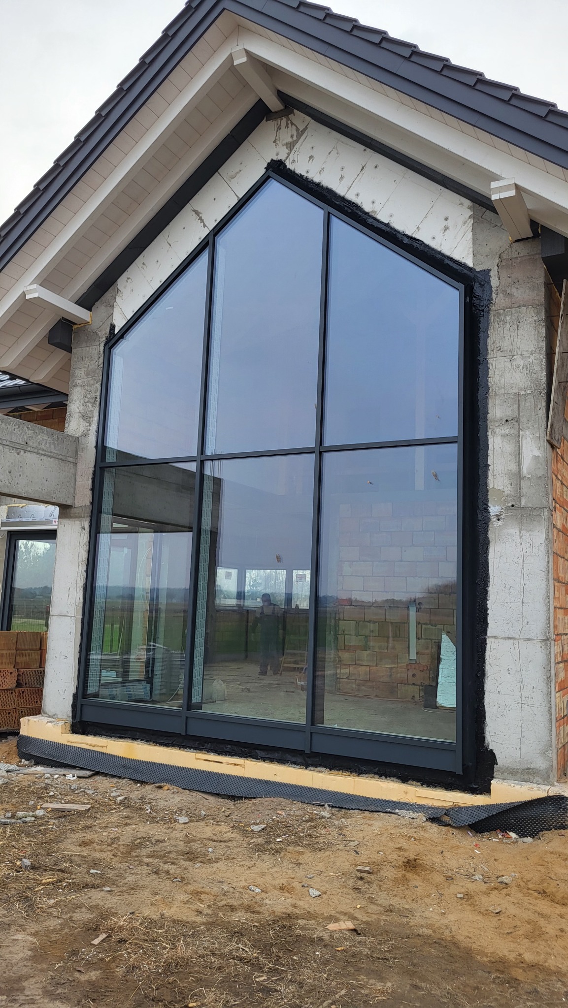 Wysokie okno aluminiowe w kształcie trapezu w surowym stanie budynku. Widoczne cegły, beton i izolacja. W oknach odbija się sylwetka osoby.