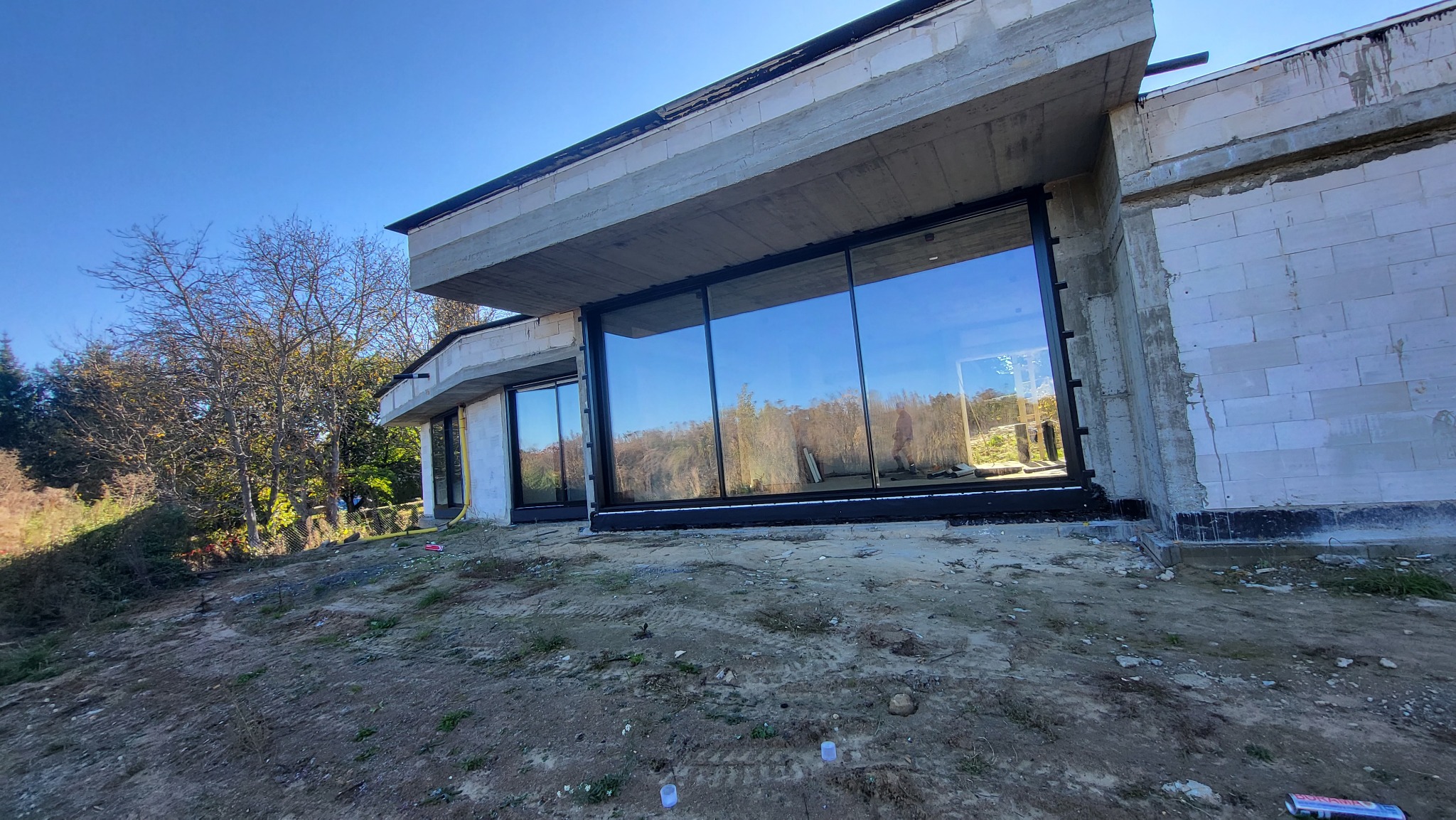 Nowoczesny dom w budowie z dużymi, panoramicznymi oknami z czarnymi ramami, odbijającymi krajobraz. Widok zewnętrzny na surowy stan budynku.