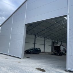 Serhii Lisetskyi SWD-BUD - Nowoczesna hala z płyt warstwowych w odcieniach szarości, z widoczną konstrukcją stalową wewnątrz. Na posadzce zaparkowane samochody.