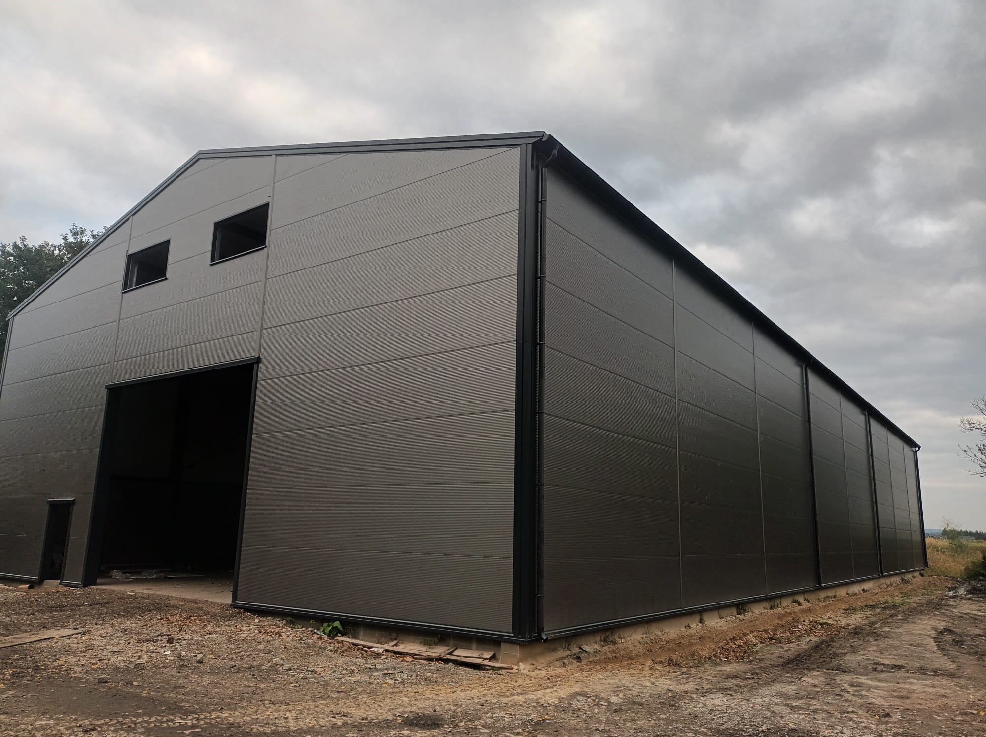 Nowoczesna hala z płyt warstwowych w odcieniach szarości, z dwoma oknami i otwartą bramą wjazdową. Widok z zewnątrz, pochmurne niebo w tle.