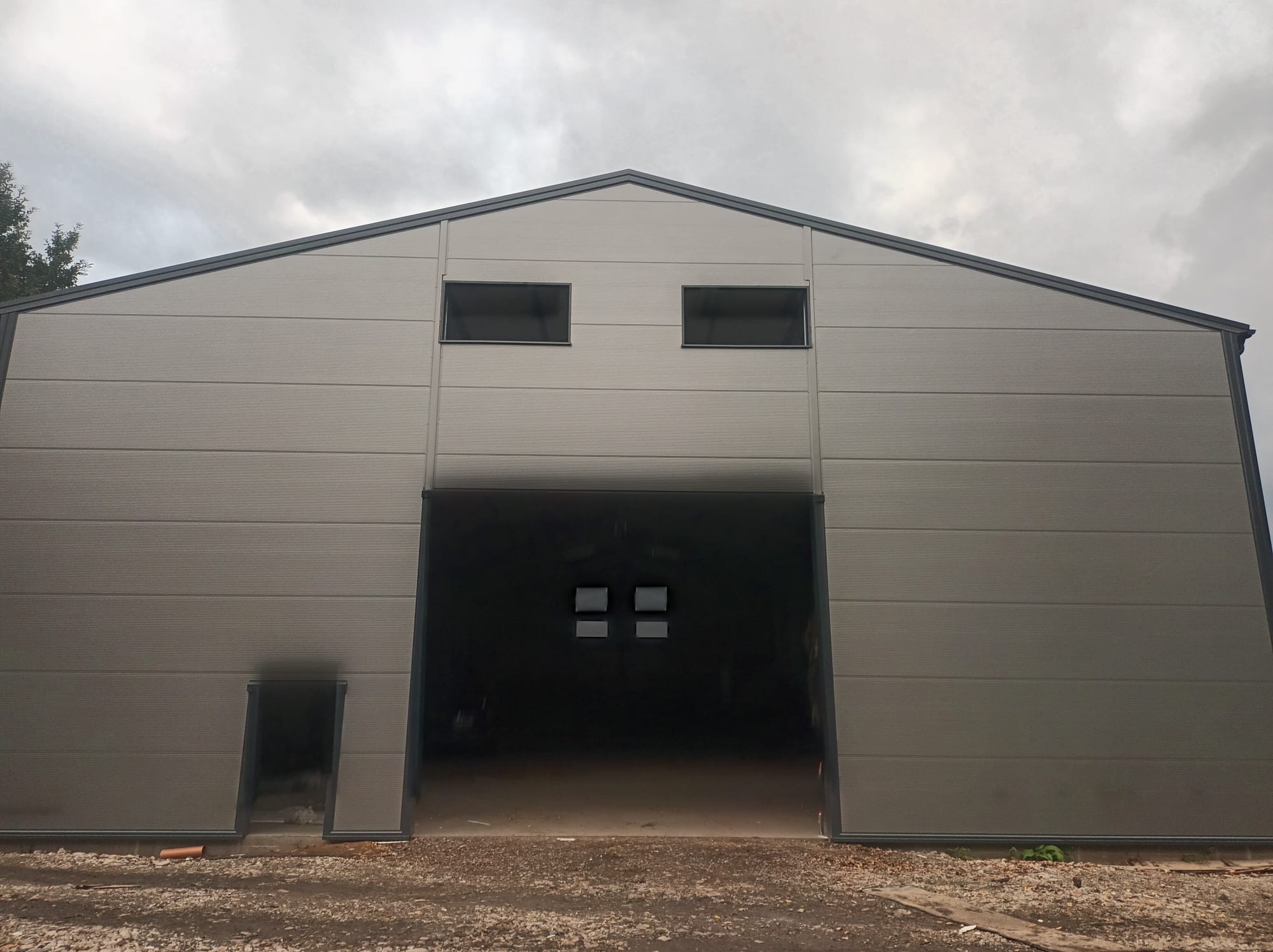 Szara hala z płyty warstwowej z otwartą bramą i małymi oknami pod dachem, widok z zewnątrz na tle pochmurnego nieba. Małe drzwi po lewej stronie.