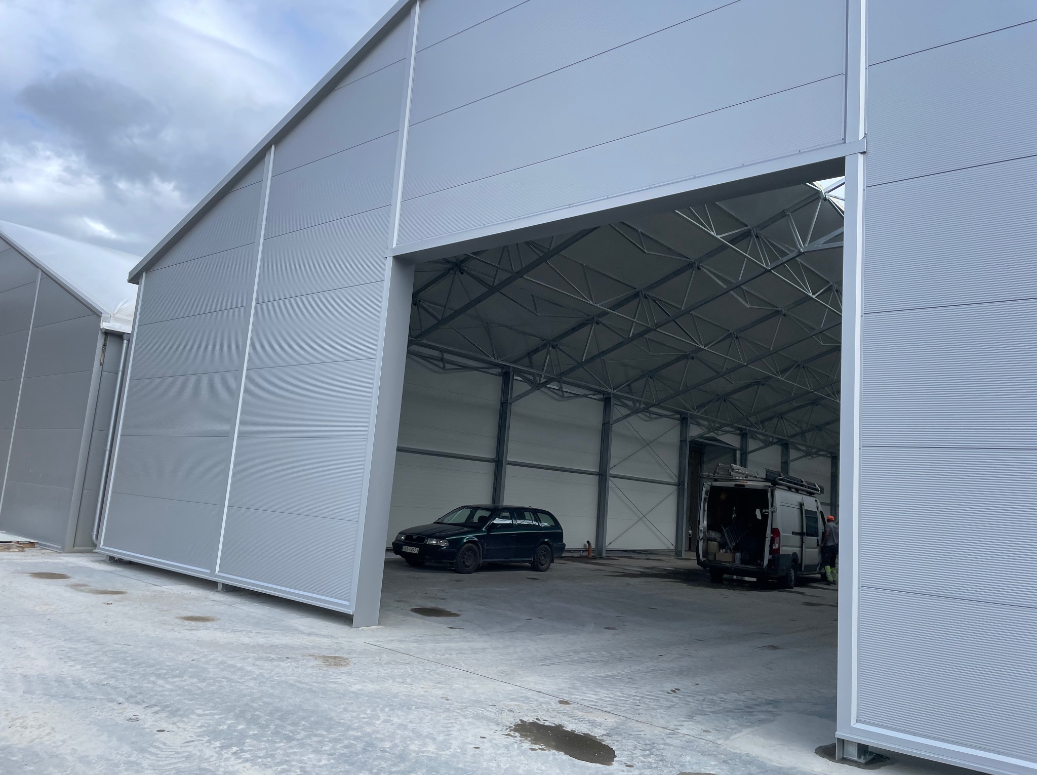 Nowoczesna hala z płyt warstwowych w odcieniach szarości, z widoczną konstrukcją stalową wewnątrz. Na posadzce zaparkowane samochody.