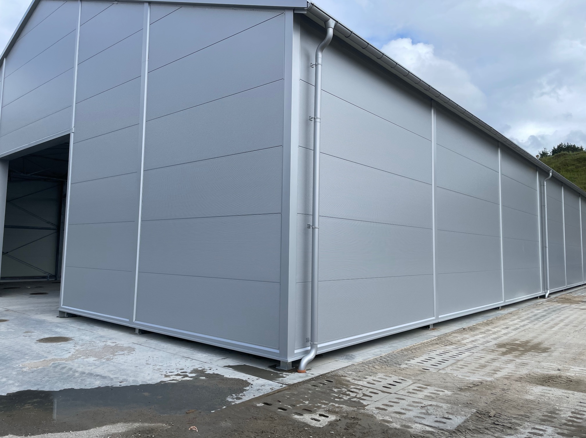 Szara hala z płyt warstwowych, widok z zewnątrz. Widoczne ryflowane panele, rynny i betonowa posadzka. Częściowo widoczne niebo i zieleń w tle.