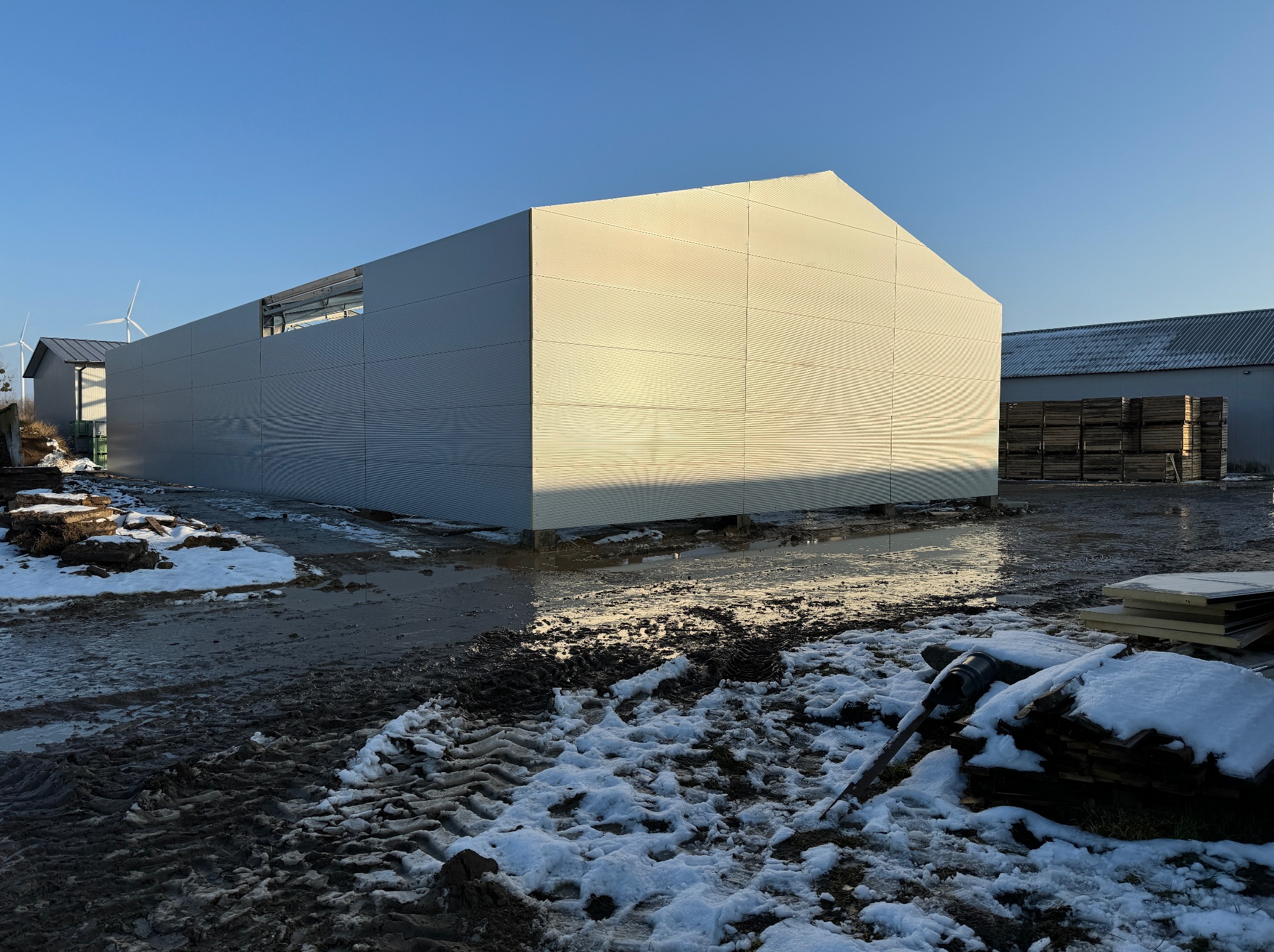 Hala w budowie, widoczne płyty warstwowe na elewacji, teren budowy z błotem i śniegiem, w tle turbiny wiatrowe. Częściowo ukończona konstrukcja.