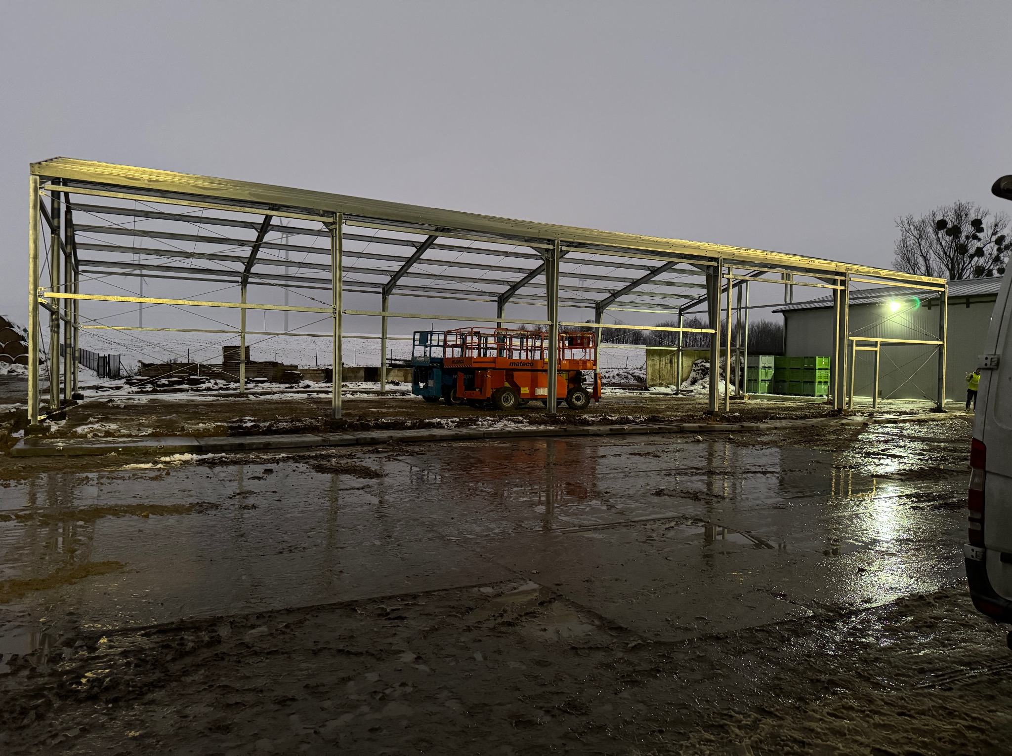 Konstrukcja stalowa hali w trakcie budowy, widoczny podnośnik koszowy i błotniste podłoże. Szara kolorystyka dnia.