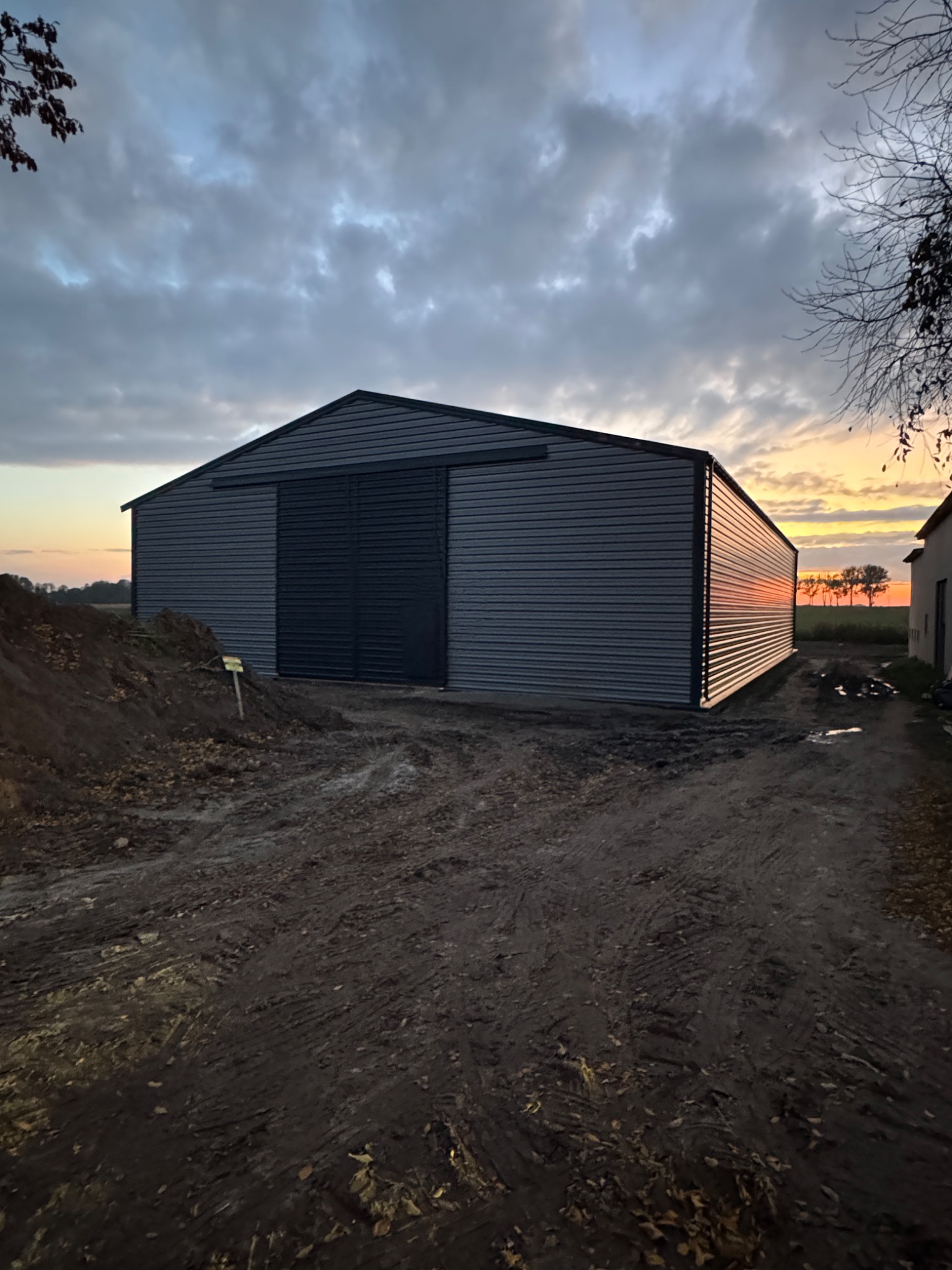 Szara hala z płyt warstwowych z przesuwnymi drzwiami na tle zachodzącego słońca. Widoczne ślady opon na ziemi i mały budynek w oddali.