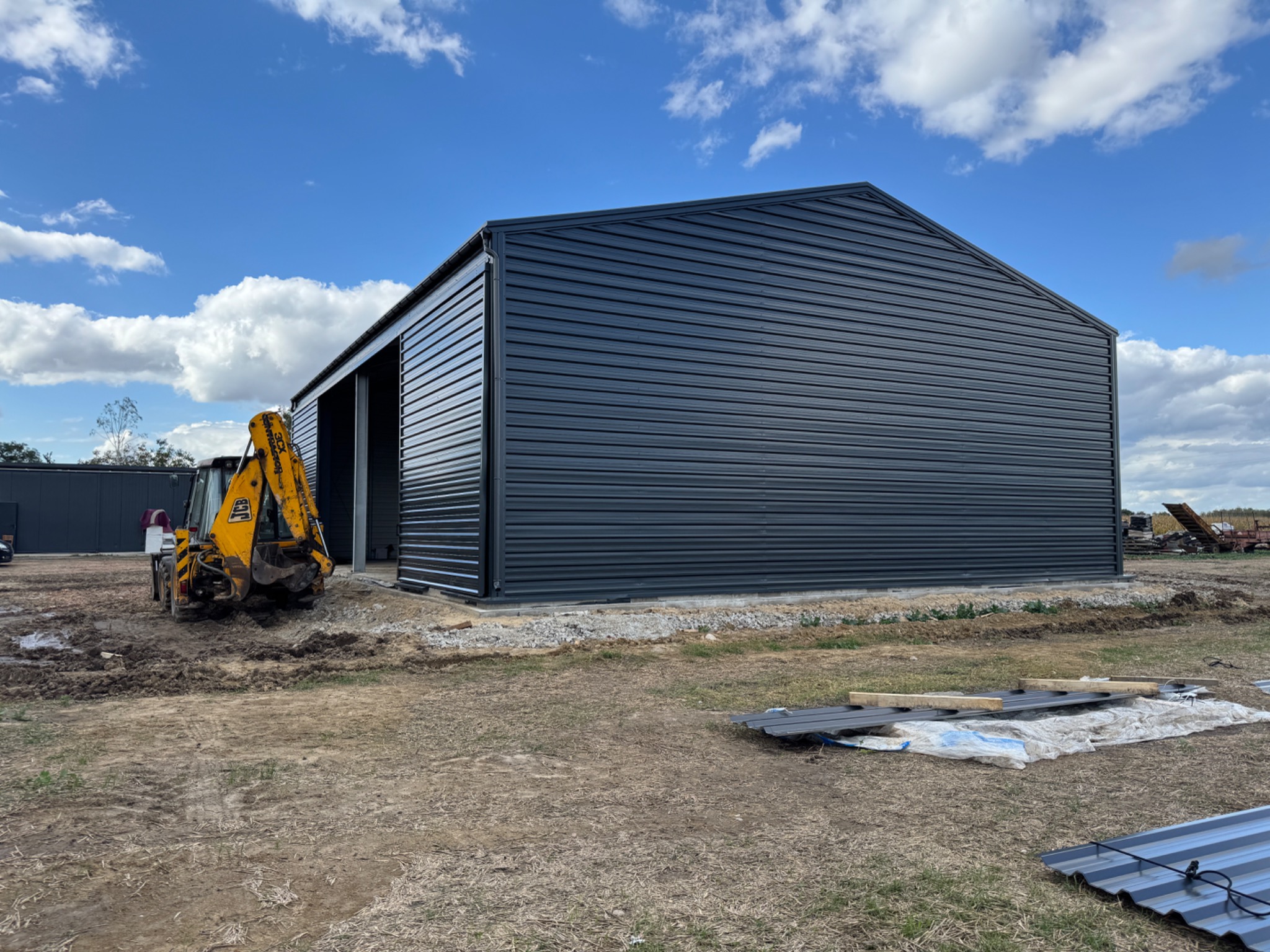 Nowoczesna hala z płyt warstwowych w kolorze grafitowym, z żółtą koparką JCB obok, na tle błękitnego nieba z chmurami. Widoczne elementy konstrukcyjne i teren budowy.