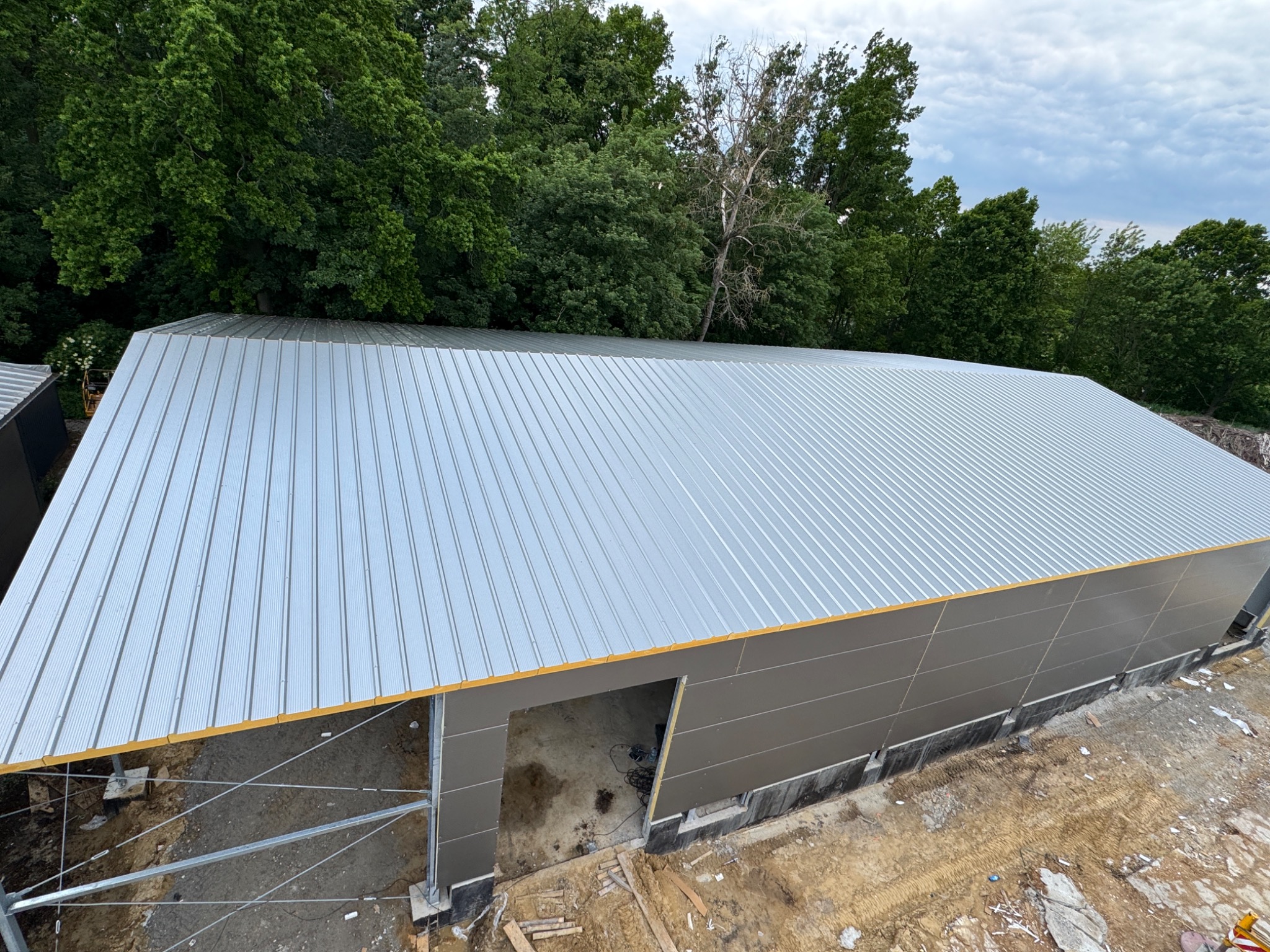 Hala w trakcie budowy z szarej płyty warstwowej, widok z góry na dach i fragment ściany, teren budowy z ziemią i zielenią w tle.