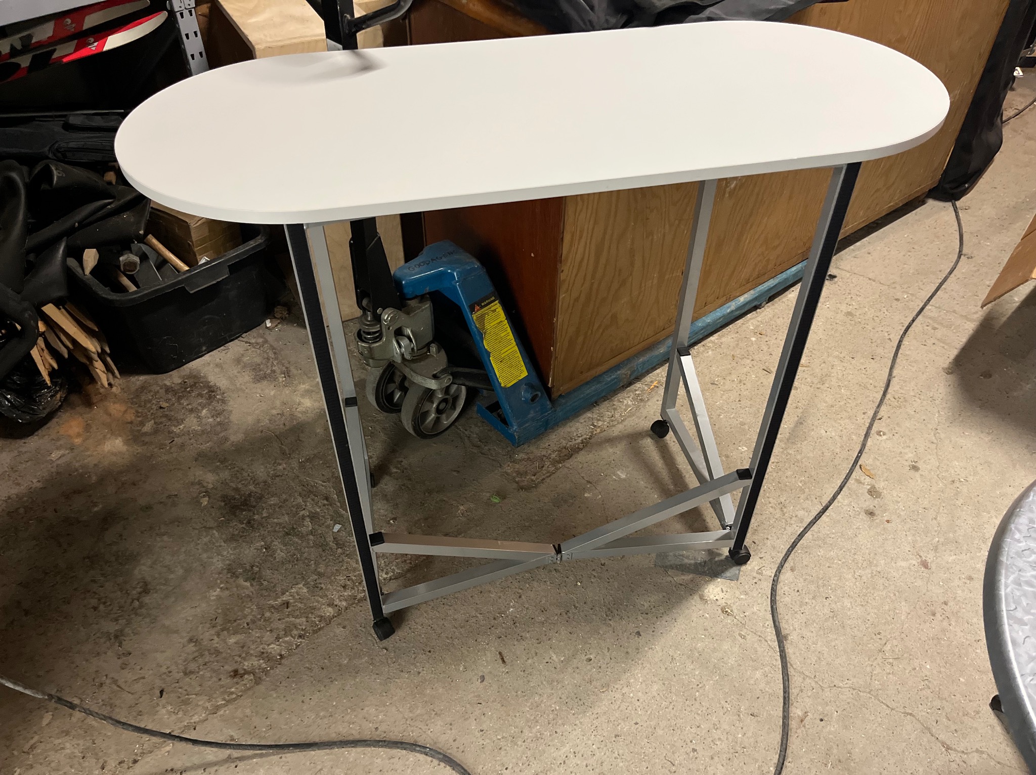 Biały, owalny stół na metalowym stelażu, z paleciakiem w tle. Stół ustawiony na betonowej podłodze, widoczne narzędzia i akcesoria w tle. Ujęcie z niskiego kąta.