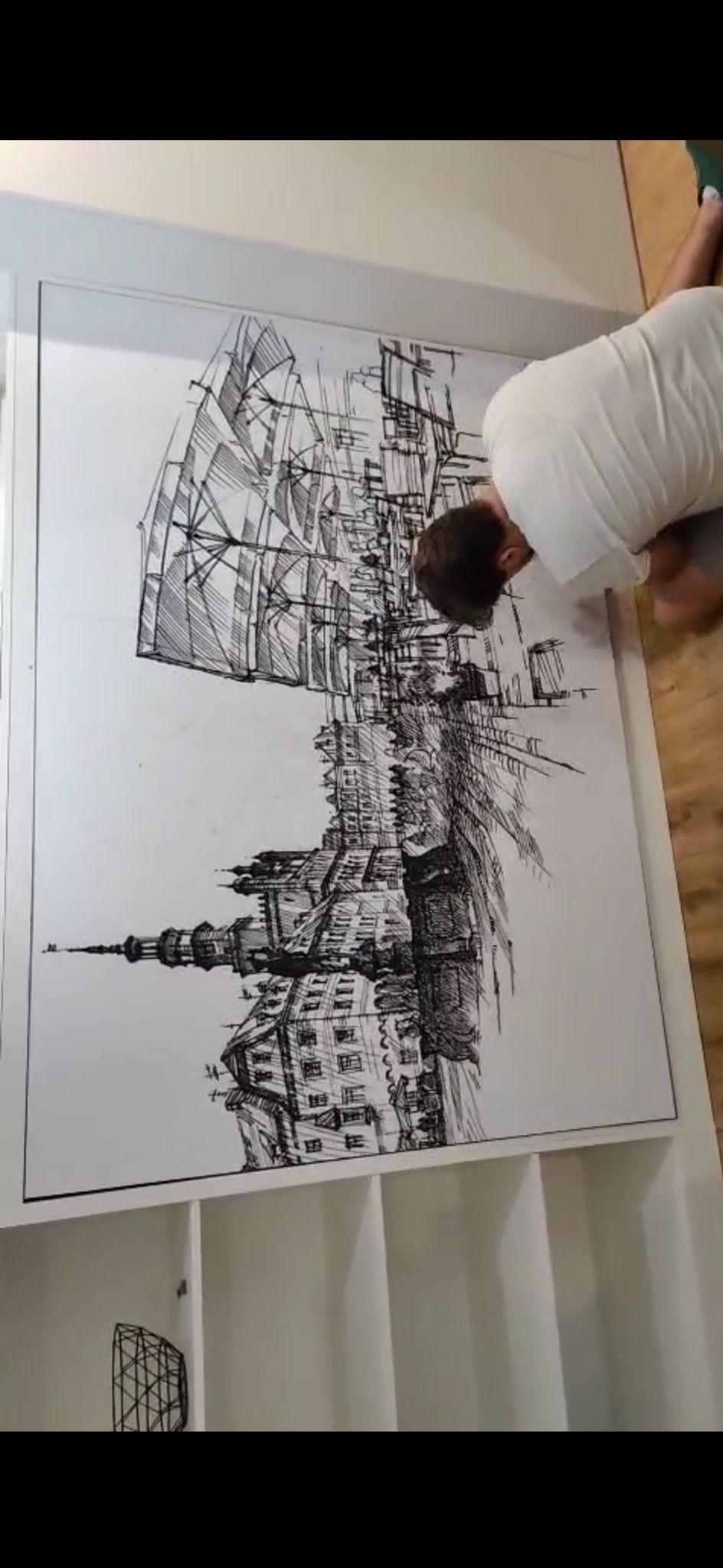 Artysta pochylony nad dużym rysunkiem tuszem przedstawiającym architekturę miejską z elementami żaglowca, umieszczonym na białym regale.