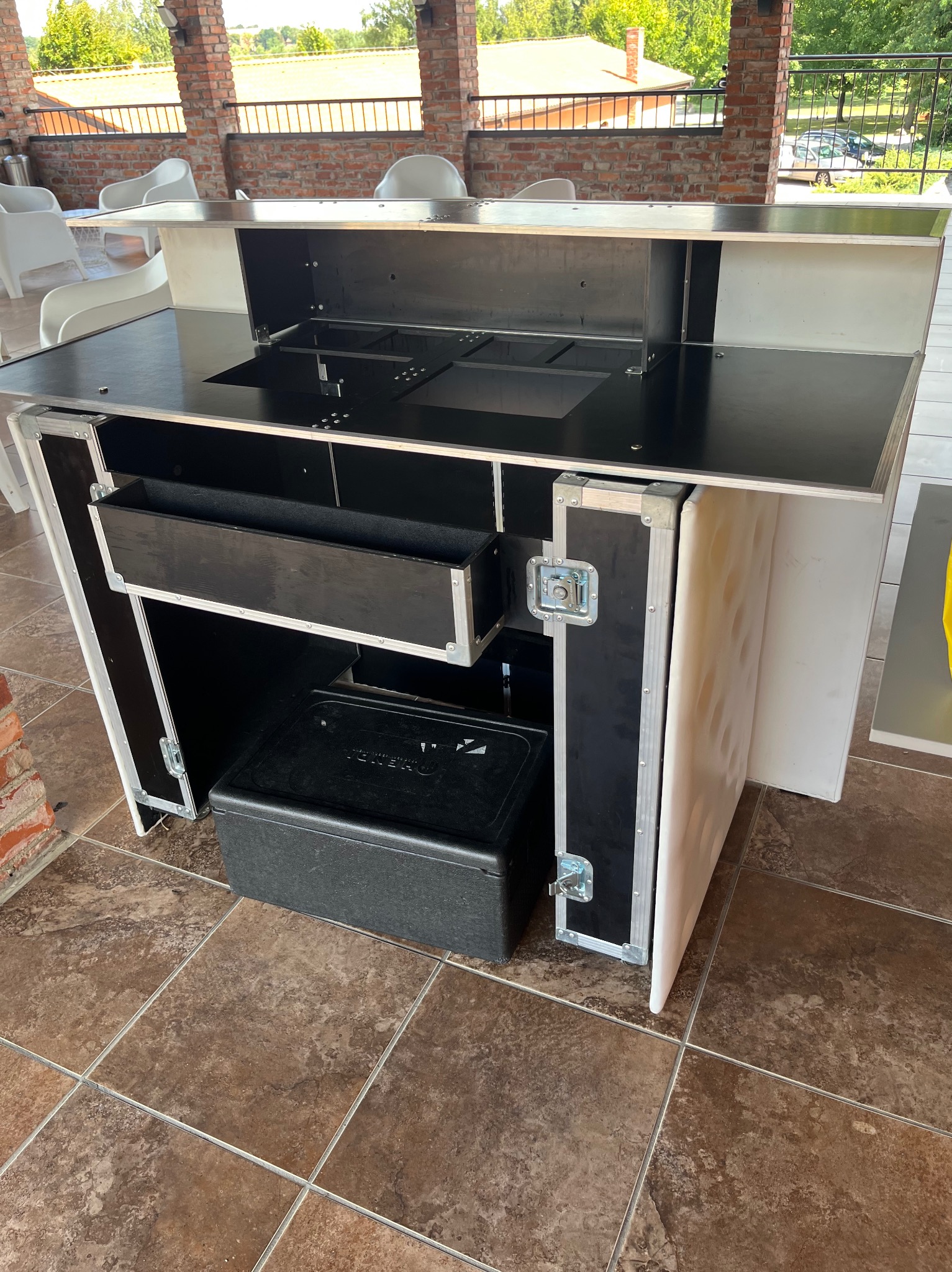 Mobilny bar cateringowy z czarnym blatem i srebrnymi okuciami, ustawiony na brązowej terakocie w zadaszonej przestrzeni. Widoczny pojemnik chłodzący.