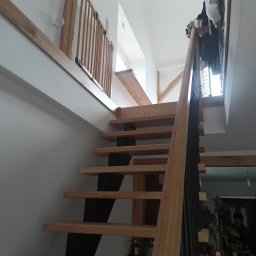 Usługi ogólnobudowlane - Drewniane schody z czarnymi wspornikami, widok z dołu, w jasnym wnętrzu z elementami drewnianymi. Balustrada z pionowych szczebli.