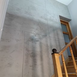 Usługi ogólnobudowlane - Ściana wykończona szarą, dekoracyjną płytą imitującą beton, widoczna z perspektywy schodów. Element wykończenia wnętrza z drewnianą balustradą.
