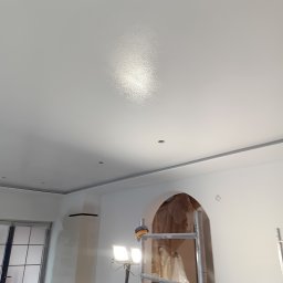 Usługi ogólnobudowlane - Gładko pomalowany sufit w Bartoszycach, widoczne oświetlenie robocze i rusztowanie. Świeżo wykończone wnętrze z łukowym przejściem w tle.