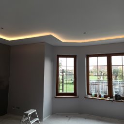 Usługi ogólnobudowlane - Pokój po malowaniu ścian na szaro, z oknami i podświetlanym sufitem. Widoczna drabina malarska i ślady prac wykończeniowych na podłodze.