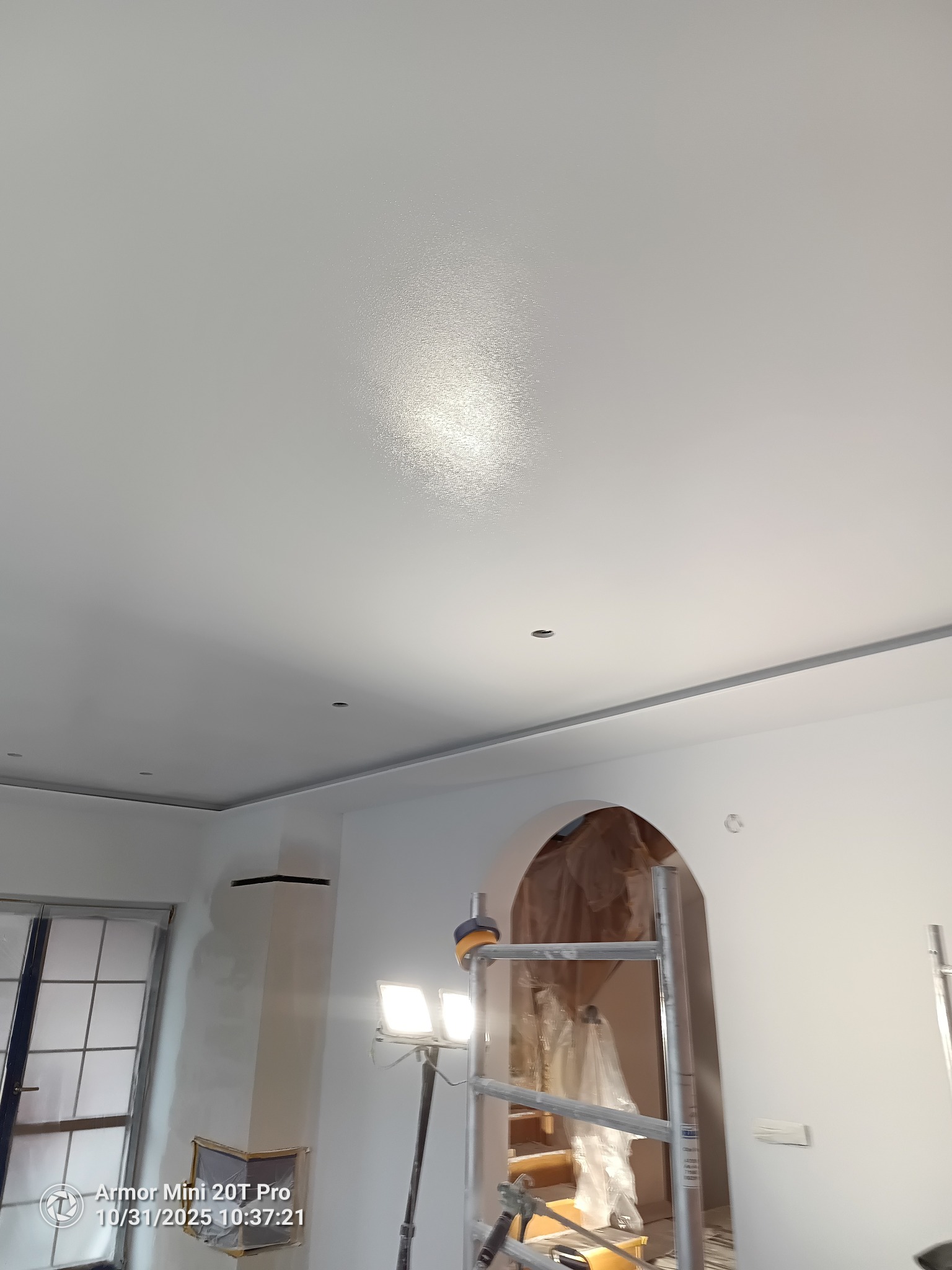 Gładko pomalowany sufit w Bartoszycach, widoczne oświetlenie robocze i rusztowanie. Świeżo wykończone wnętrze z łukowym przejściem w tle.