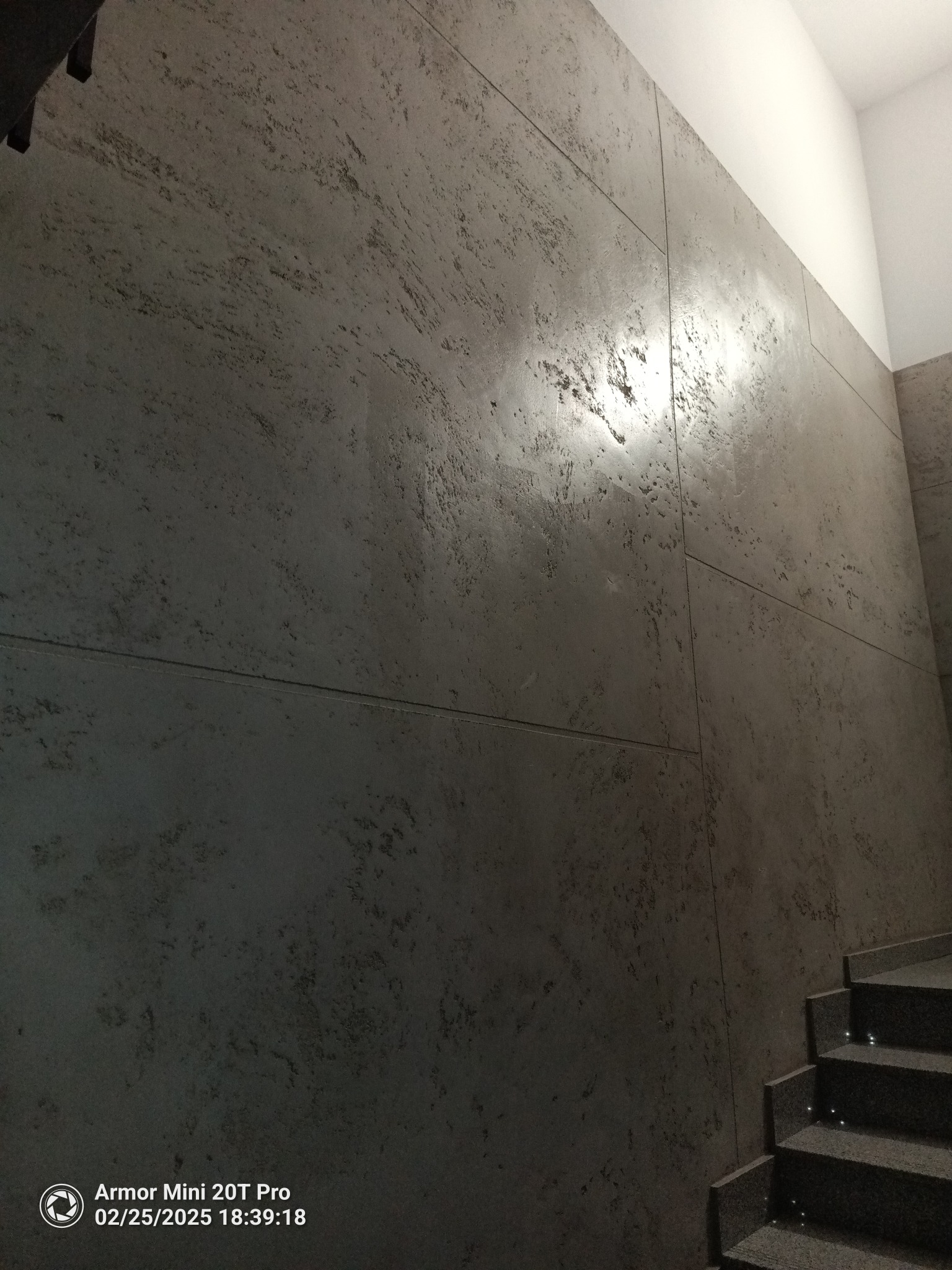 Ściana wykończona dekoracyjną, szarą strukturą przypominającą kamień, podzielona na panele. Widoczne schody z podświetleniem. Minimalistyczny design.