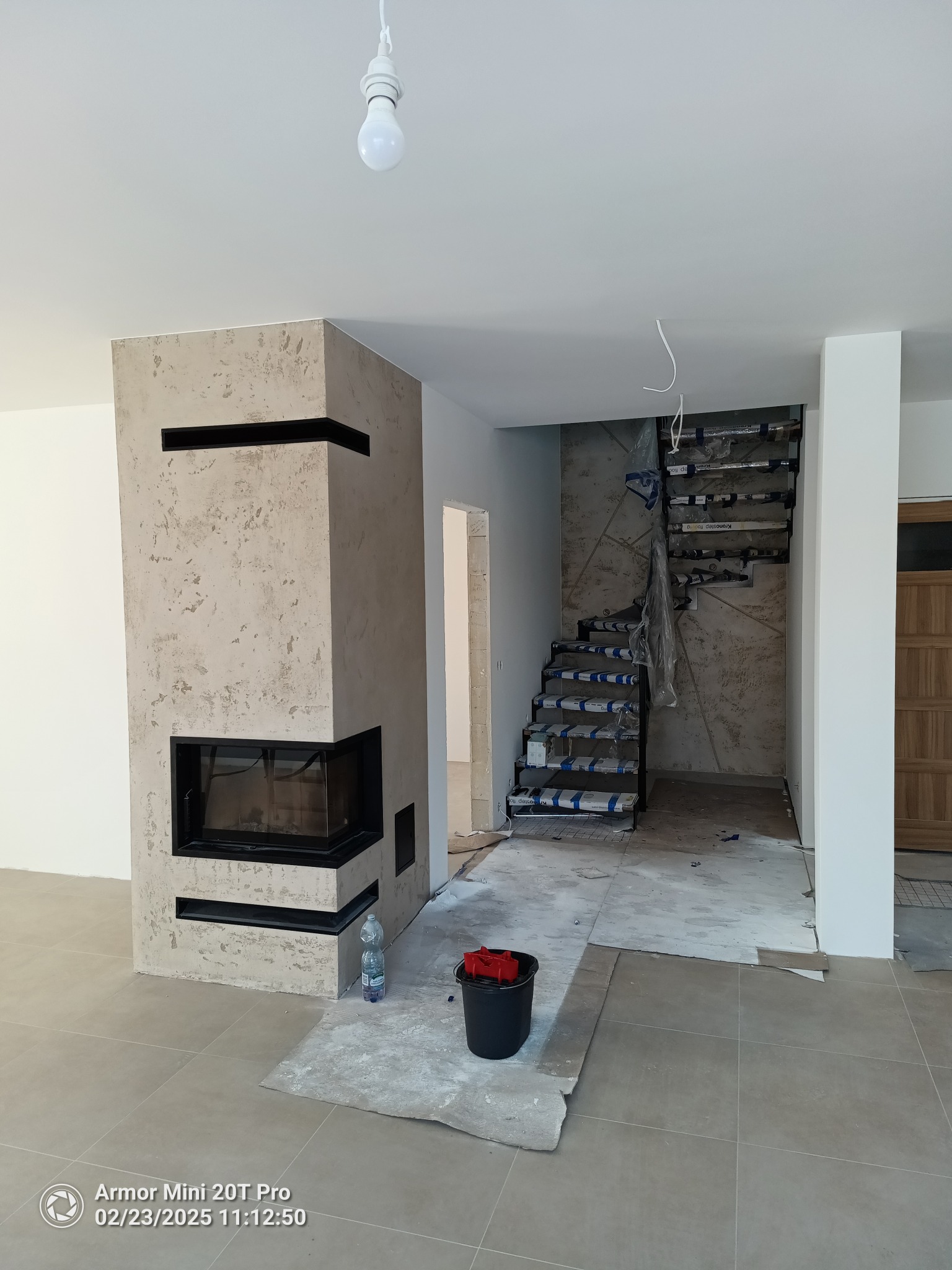 Wnętrze w trakcie wykańczania: kominek z dekoracyjnym tynkiem, schody z zabezpieczeniami, surowa podłoga. Widoczne materiały budowlane i narzędzia.