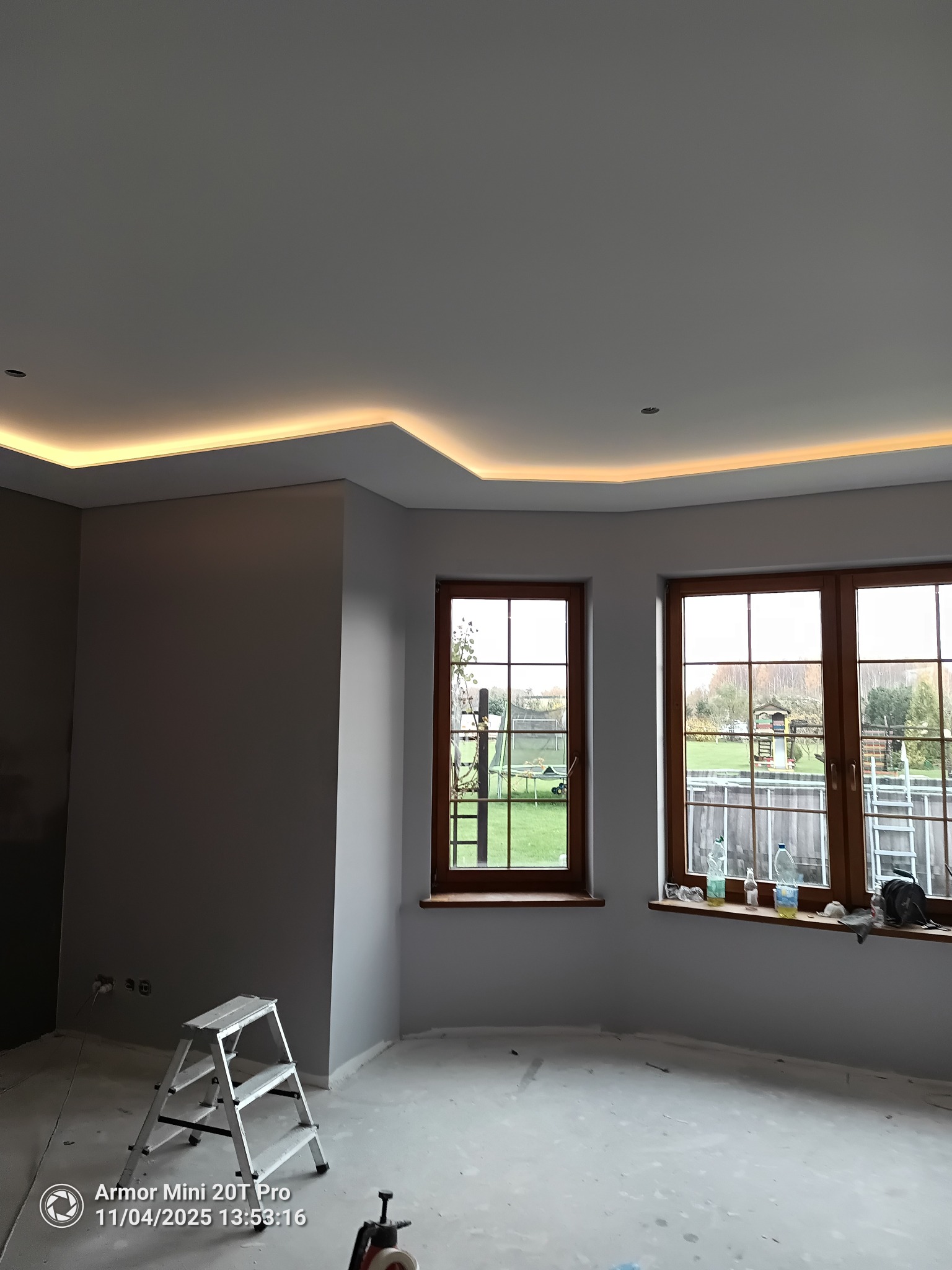 Pokój po malowaniu ścian na szaro, z oknami i podświetlanym sufitem. Widoczna drabina malarska i ślady prac wykończeniowych na podłodze.