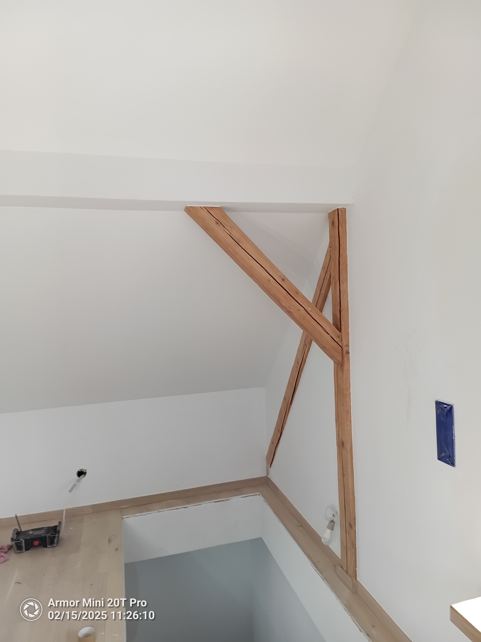 Białe wnętrze poddasza z drewnianą konstrukcją nośną, widoczne narzędzia malarskie i zabezpieczone elementy. Świeżo pomalowane ściany i sufit.