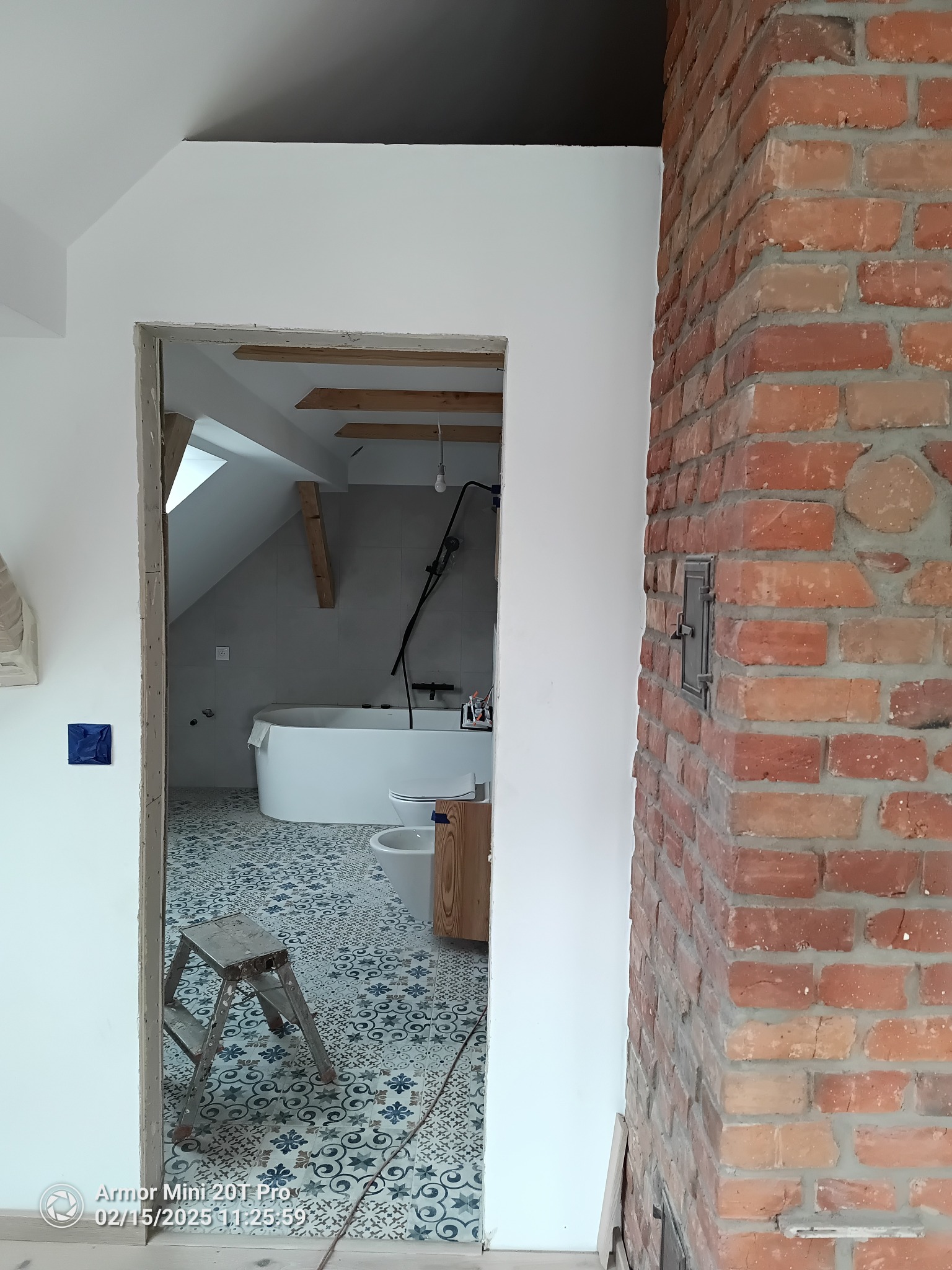 Wnętrze łazienki w trakcie remontu na poddaszu: widoczna wanna, toaleta, drabina i fragment ściany z cegły. Białe ściany kontrastują z wzorzystą podłogą.