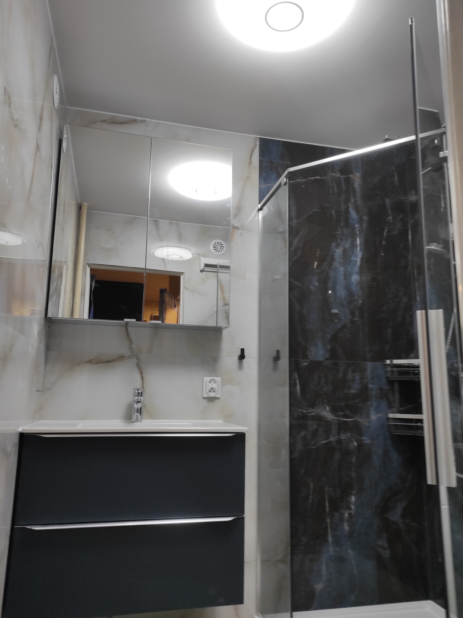 Nowoczesna łazienka z kabiną prysznicową typu walk-in, umywalką z szafką w kolorze grafitowym i lustrem w szafce. Marmurowe płytki na ścianach i sufitowe oświetlenie.