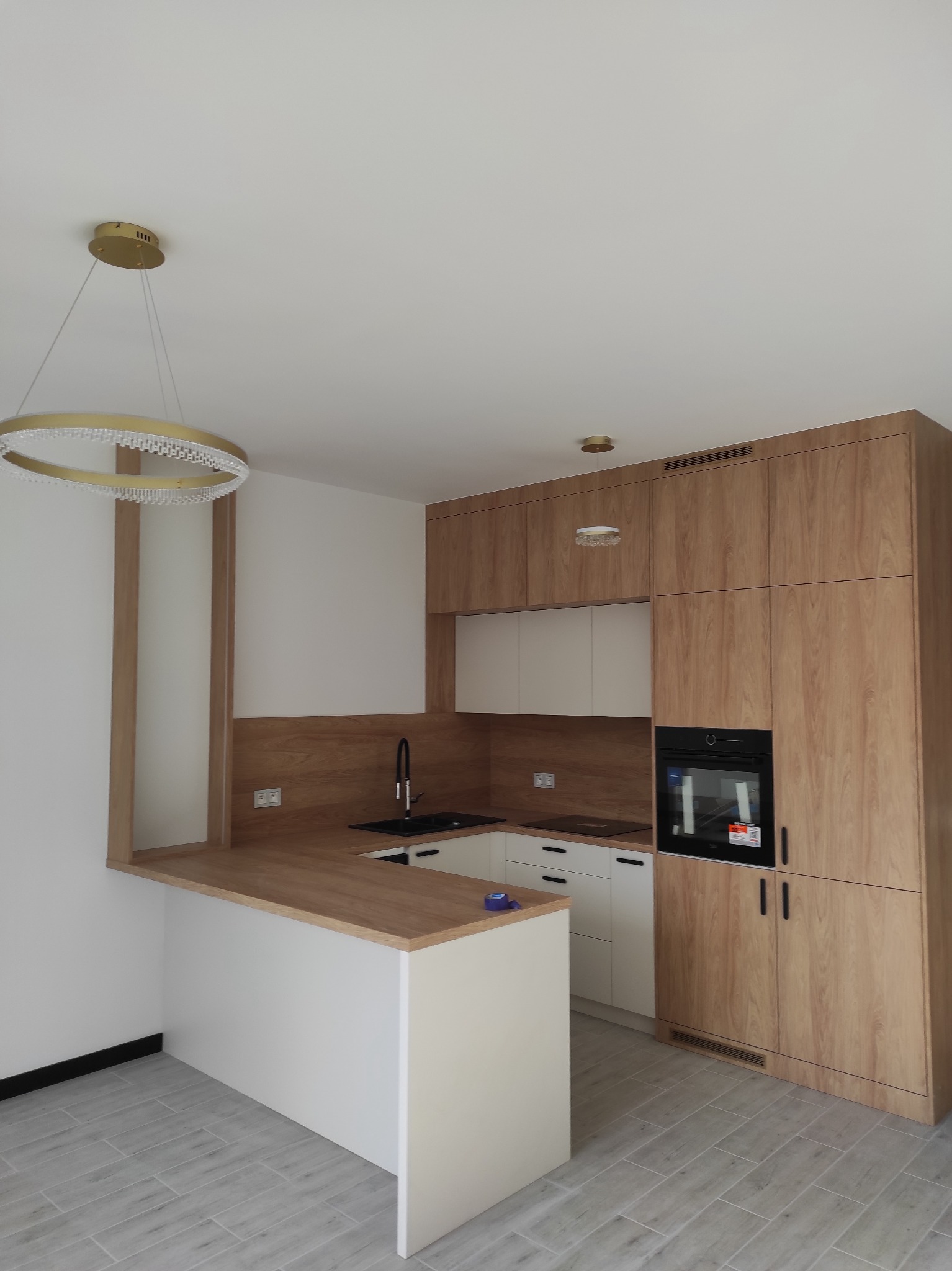 Nowoczesna kuchnia z drewnianymi szafkami, białymi frontami i wyspą. Uwagę przyciąga designerska lampa wisząca nad blatem. Jasne płytki podłogowe.