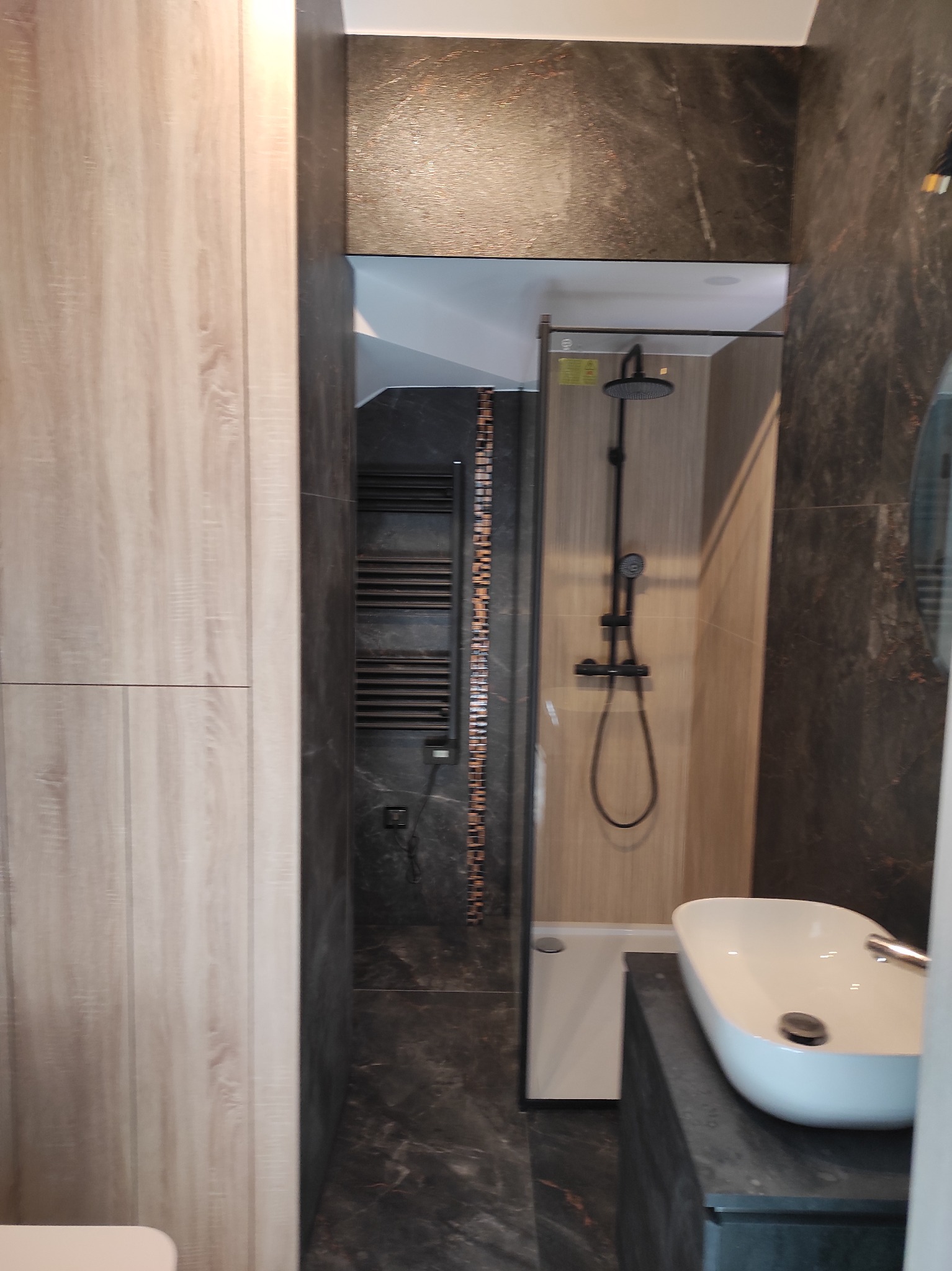 Nowoczesna łazienka z kabiną prysznicową typu walk-in, umywalką nablatową i grzejnikiem drabinkowym. Ciemne płytki imitujące kamień i drewnopodobne panele ścienne.