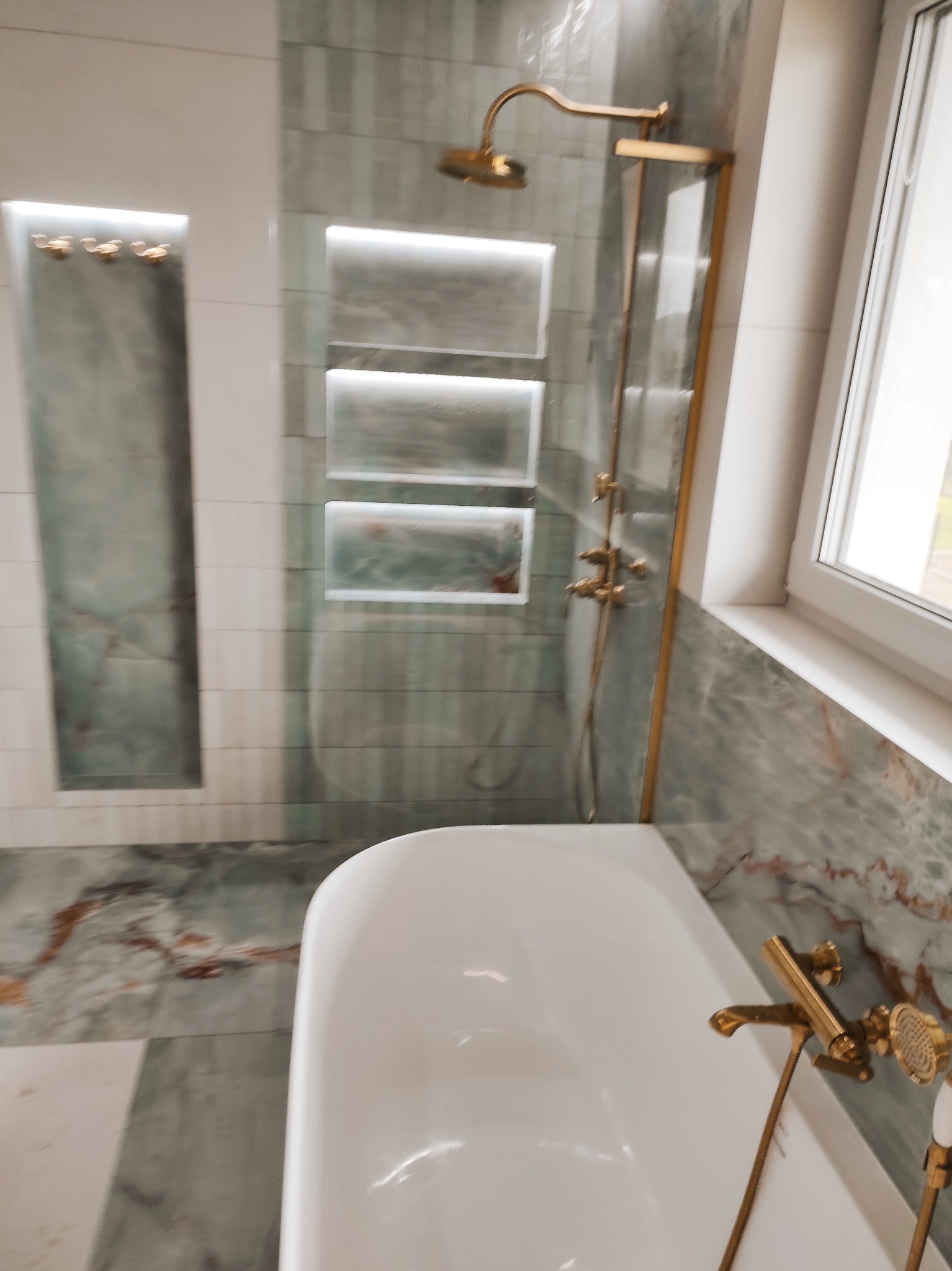 Elegancka łazienka z wanną, złotą armaturą i podświetlanymi niszami w szaro-białych płytkach imitujących marmur. Widok na okno i kabinę prysznicową.