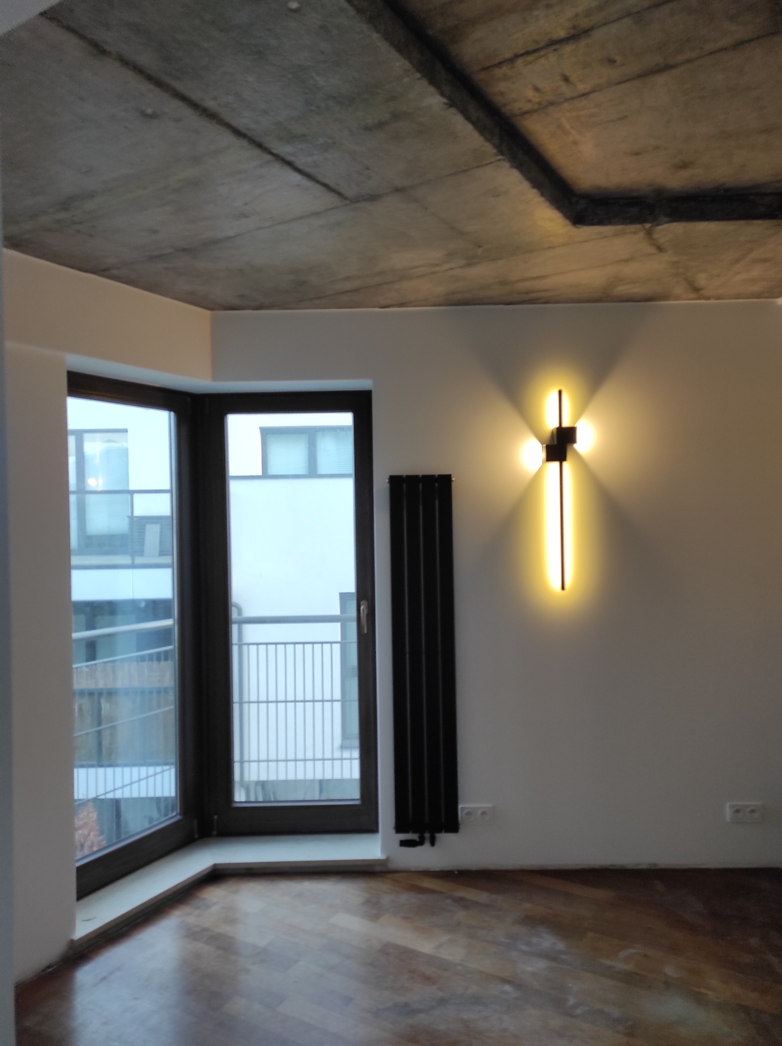 Nowoczesne wnętrze z parkietem, betonowym sufitem i designerskim oświetleniem ściennym w kształcie krzyża. Czarny grzejnik panelowy przy oknie.